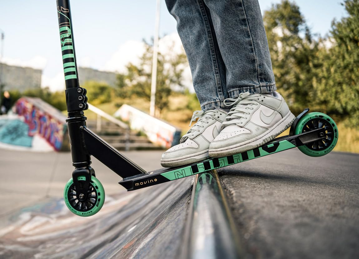 Ein Mensch in Jeans und weißen Sneakers macht einen Stunt mit einem schwarz-türkisen Stunt-Scooter auf einer Rampe.