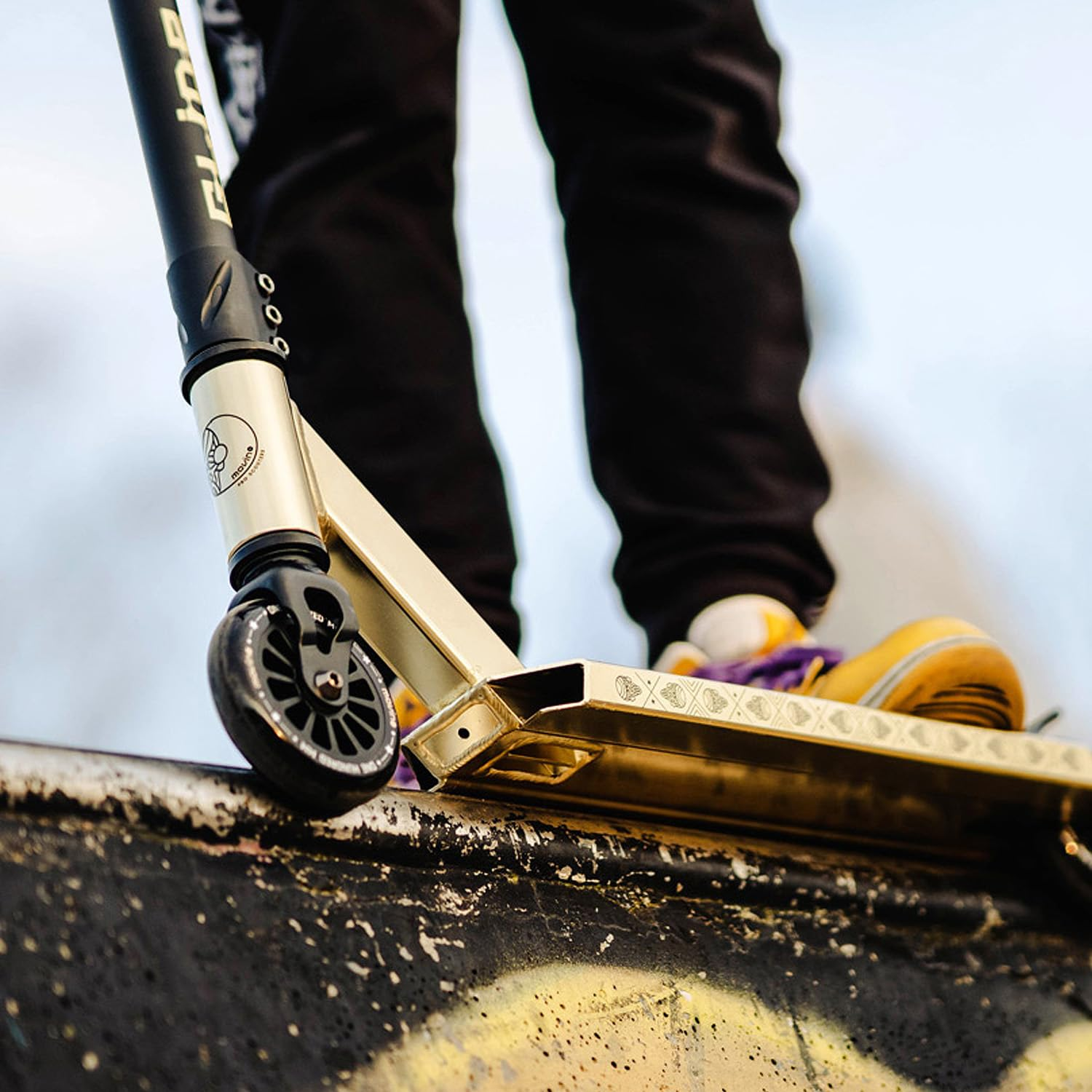 Eine Person steht auf einem goldenen Stunt-Scooter, der auf einer grauen Skatepark-Rampe positioniert ist.