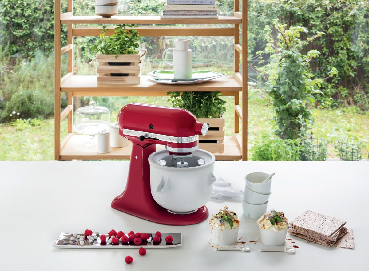 Rote Küchenmaschine mit Eisschüssel auf weißem Tisch, davor Himbeeren und Desserts, vor Holzregal.