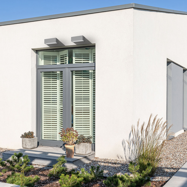 Fachada moderna de casa blanca con puerta gris, persianas, dos luces exteriores y plantas de jardín.