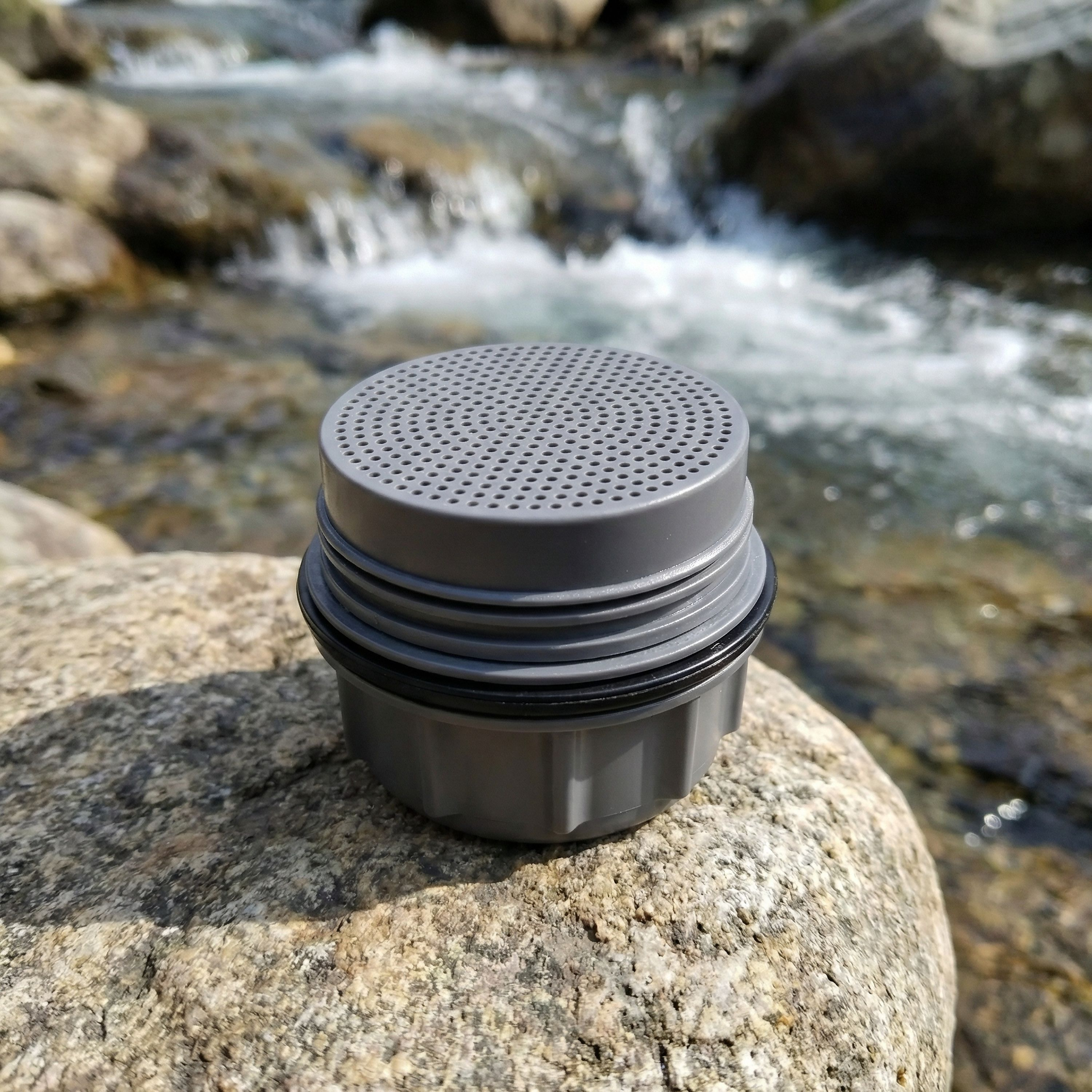 Ein grauer Wasserfilteraufsatz mit Löchern steht auf einem glatten Stein vor einem fließenden Bach.