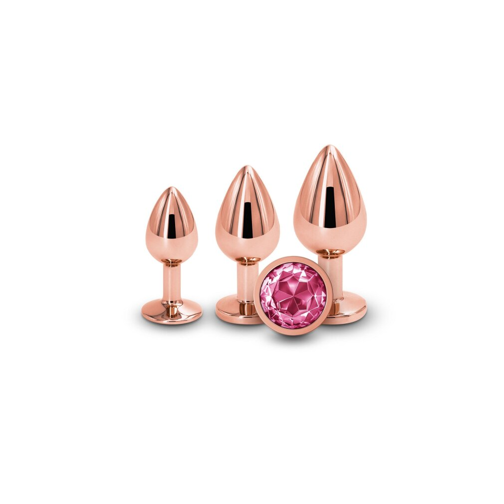 Drei roségoldene Analplugs in verschiedenen Größen mit einem facettierten rosa Schmuckstein davor.