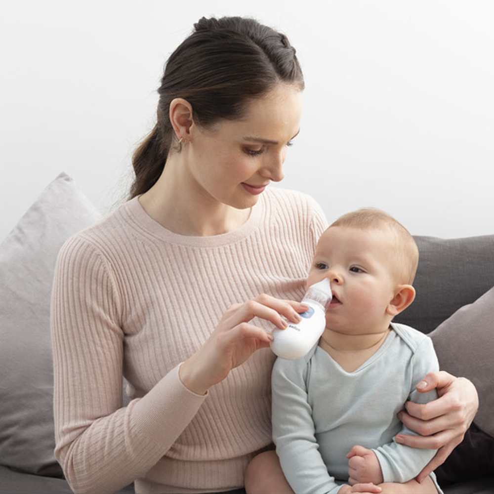 Una madre usa delicatamente un aspiratore nasale sul suo bambino su un divano grigio.