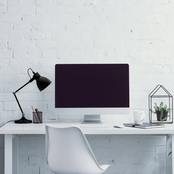 Un escritorio blanco moderno con computadora, lámpara, planta y silla blanca contra una pared de ladrillo blanco.