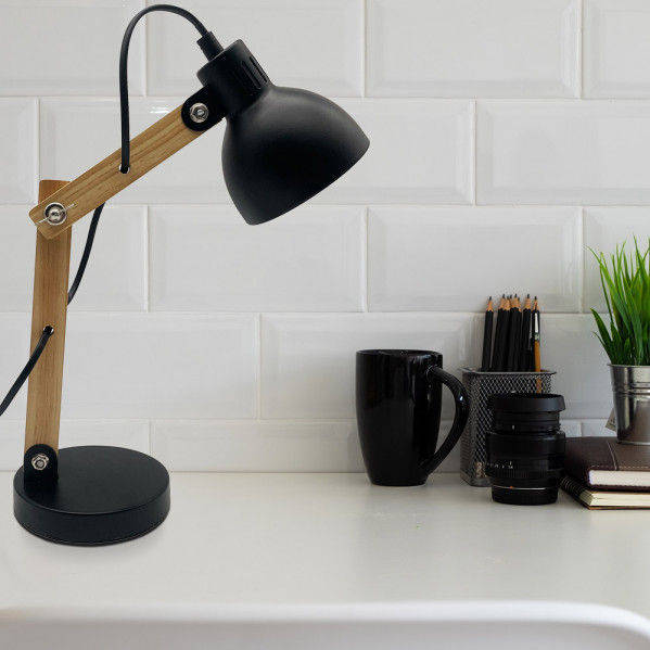 Una lámpara de escritorio negra y de madera sobre una mesa blanca frente a azulejos blancos, con una taza.