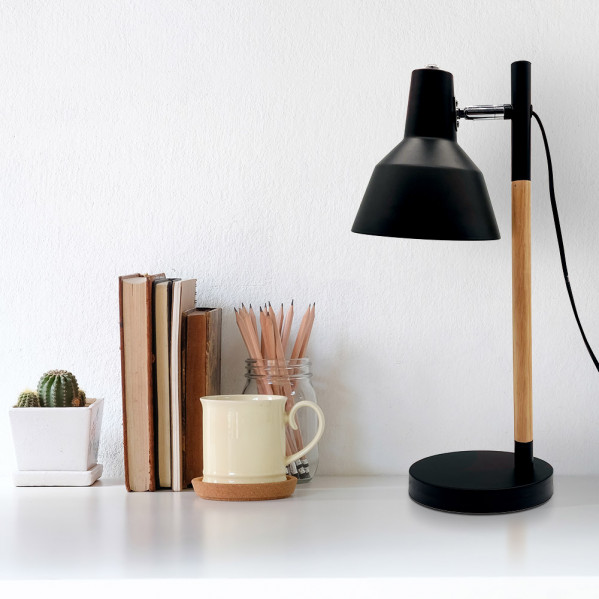 Lámpara de escritorio negra y de madera, libros, taza, tarro de lápices y un pequeño cactus contra una pared blanca.