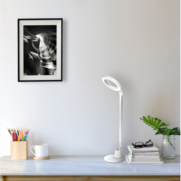 Una lámpara de escritorio LED blanca en una mesa blanca con portalápices, taza, libros y cuadro.
