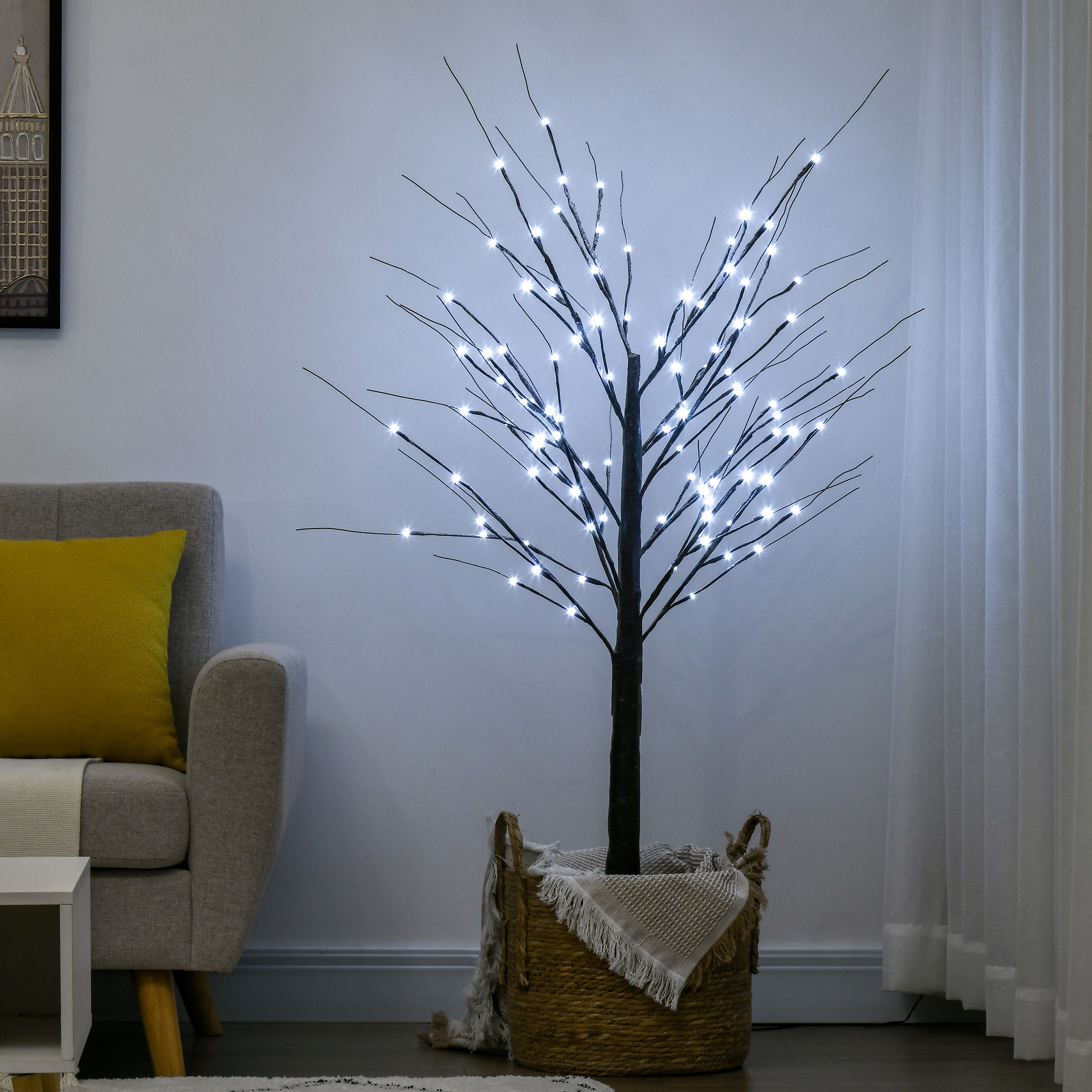 Un árbol artificial con luces LED en una cesta tejida se encuentra junto a un sofá gris con un cojín amarillo contra una pared blanca.