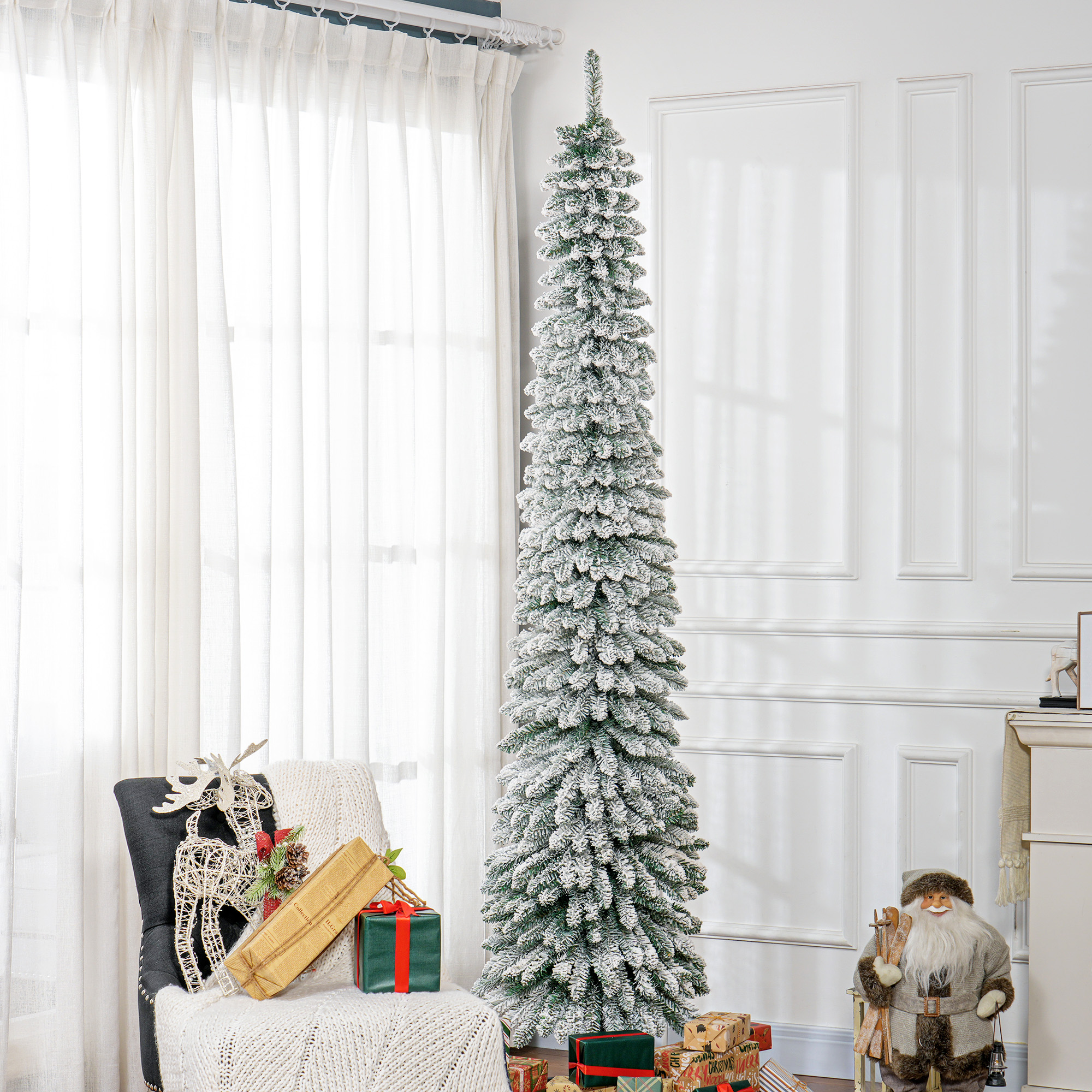Alto árbol de Navidad nevado junto a una silla con regalos, una figura de reno y una figura de Papá Noel.