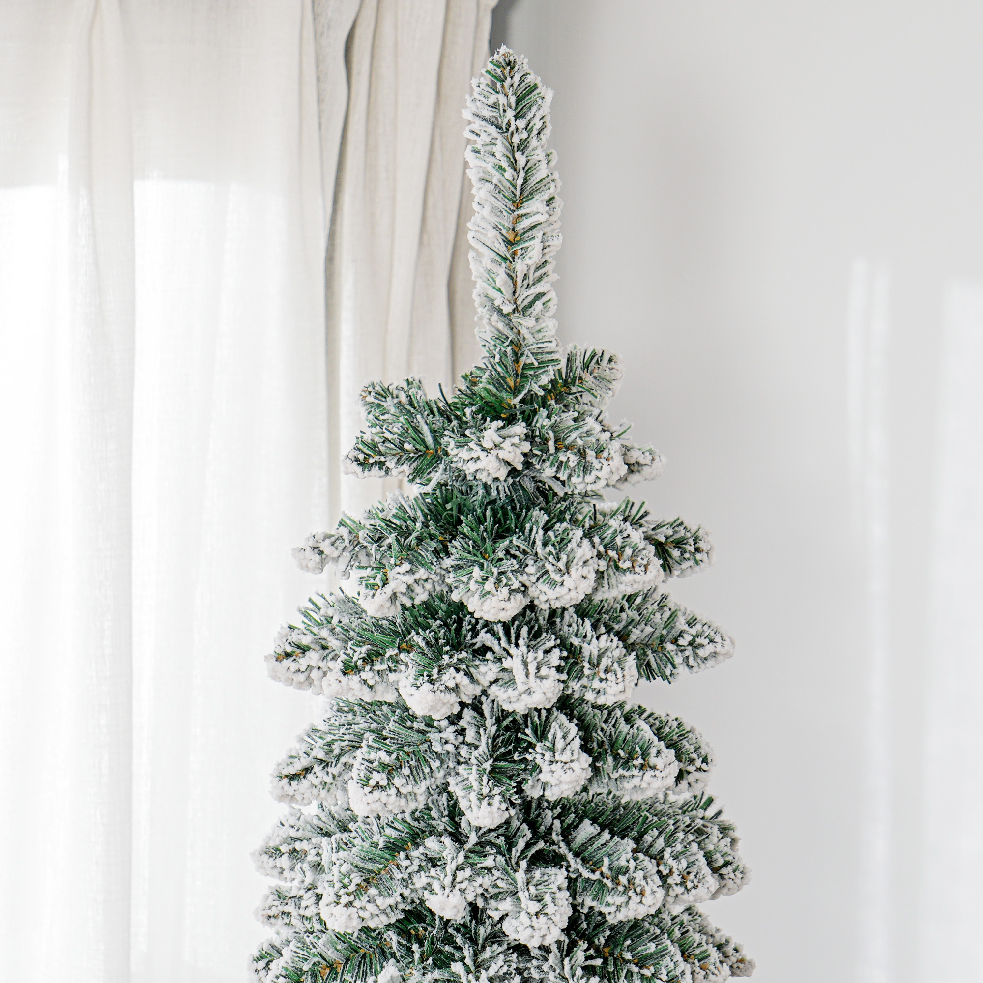 Un árbol de Navidad artificial nevado se encuentra en el interior contra una pared blanca y una cortina clara.