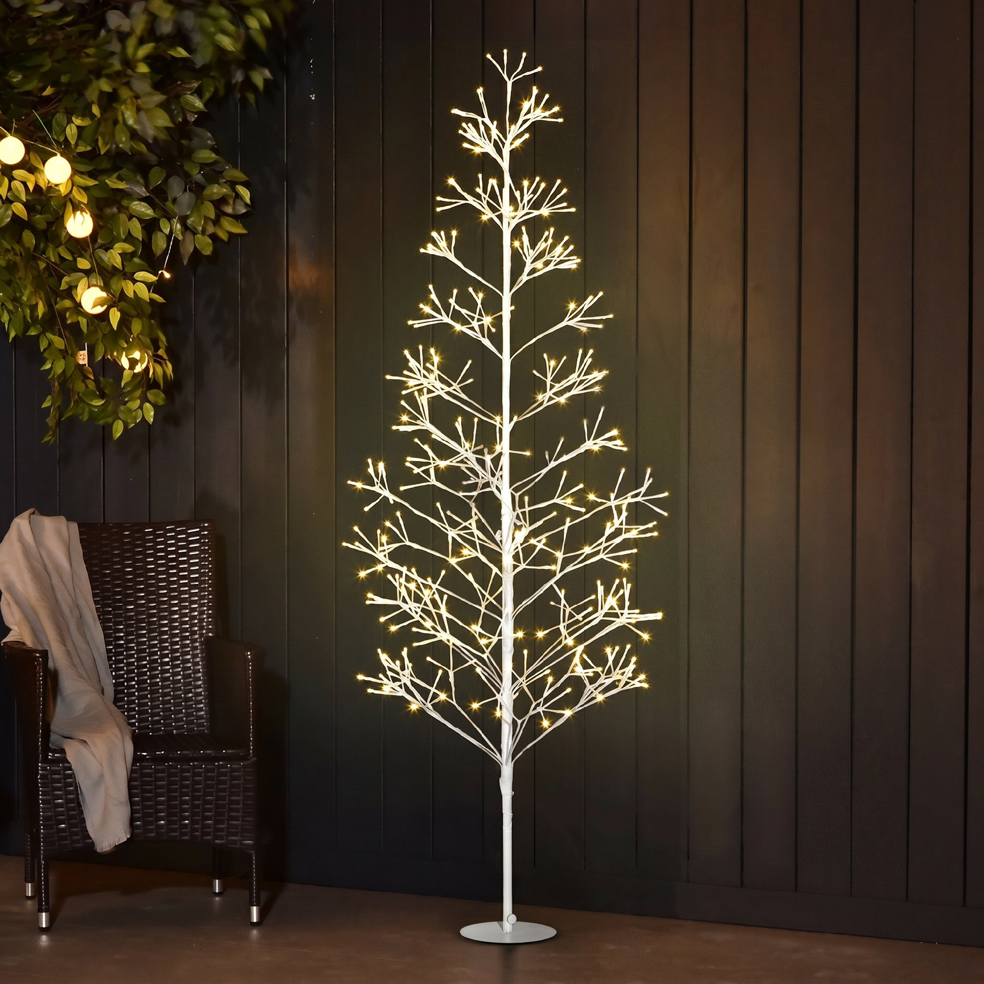 Un árbol de Navidad LED blanco y brillante se encuentra contra una pared oscura al lado de un sillón de ratán.