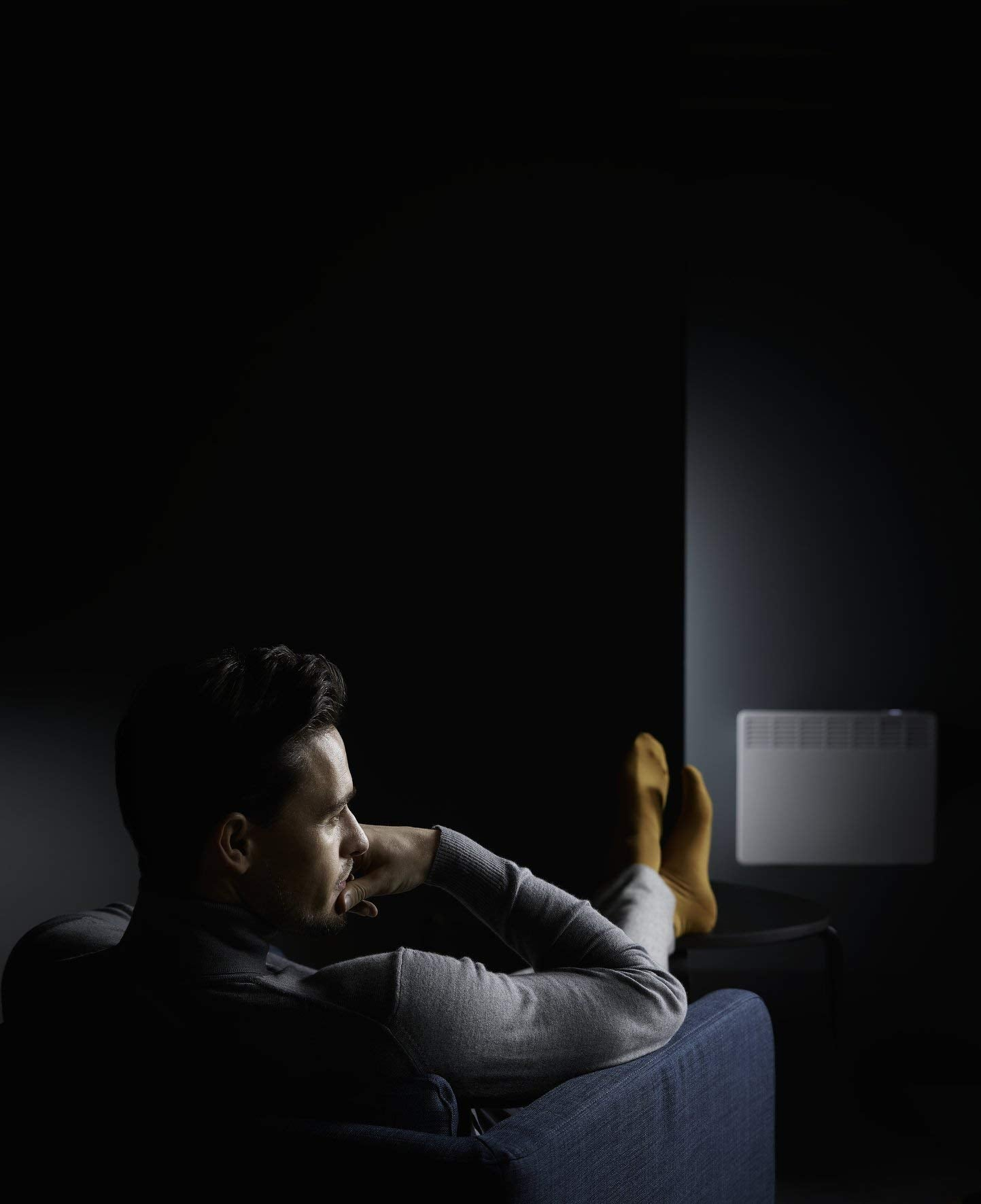 Un uomo in maglione grigio siede su un divano blu in una stanza buia, con i piedi alzati e lo sguardo rivolto a una stufa.