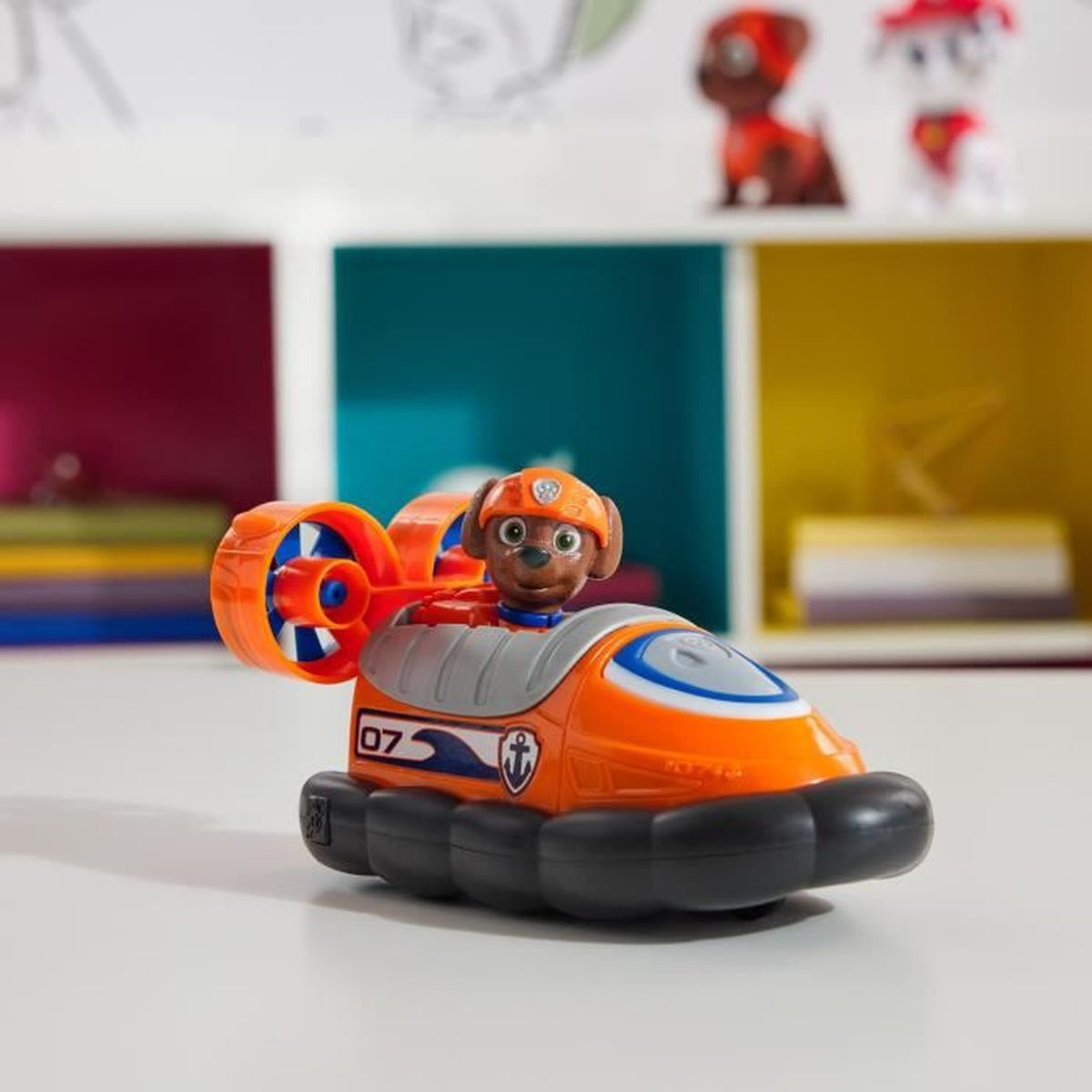 Ein orangefarbenes Paw Patrol Hovercraft-Spielzeug mit Figur vor Regalen.