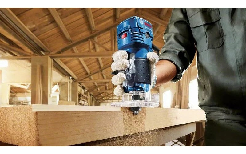 İş kıyafetli ve eldivenli bir işçi, atölyede ahşap bir kereste üzerinde mavi bir Bosch freze makinesi kullanıyor.