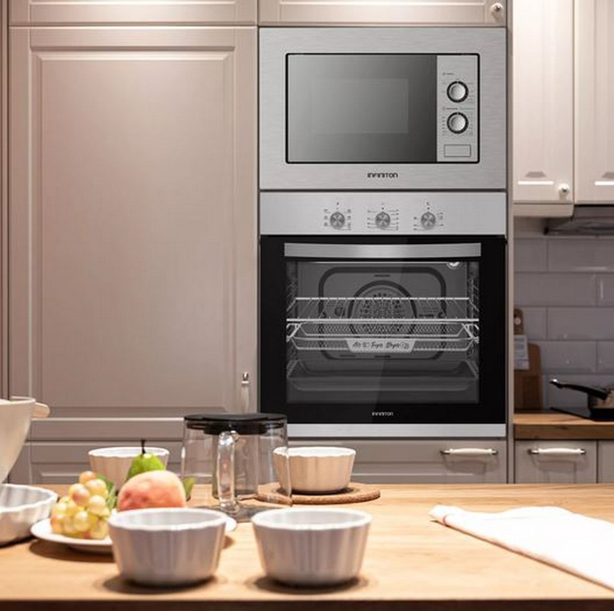 Un horno y microondas plateados empotrados en una cocina luminosa con una encimera de madera con cuencos y fruta.