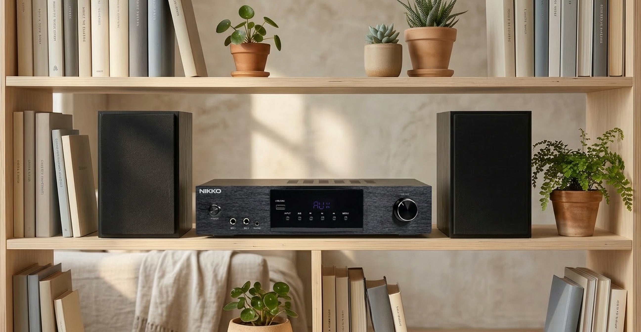 Ein schwarzer Nikko-Audio-Receiver zwischen zwei schwarzen Lautsprechern auf einem Holzbreg mit Büchern und Topfpflanzen.
