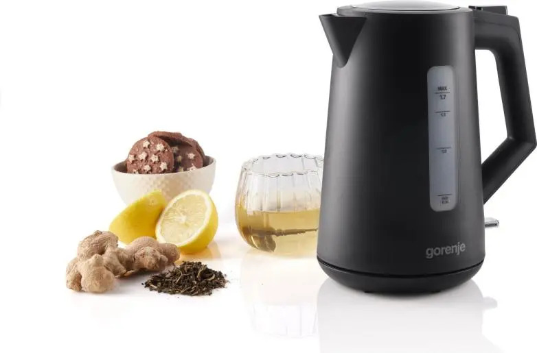 Bollitore elettrico nero accanto a tazza da tè, biscotti, zenzero, limone e foglie di tè su sfondo bianco.