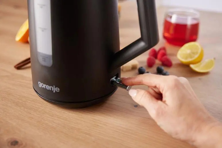 Una mano accende un bollitore elettrico nero Gorenje su un tavolo di legno con frutta.