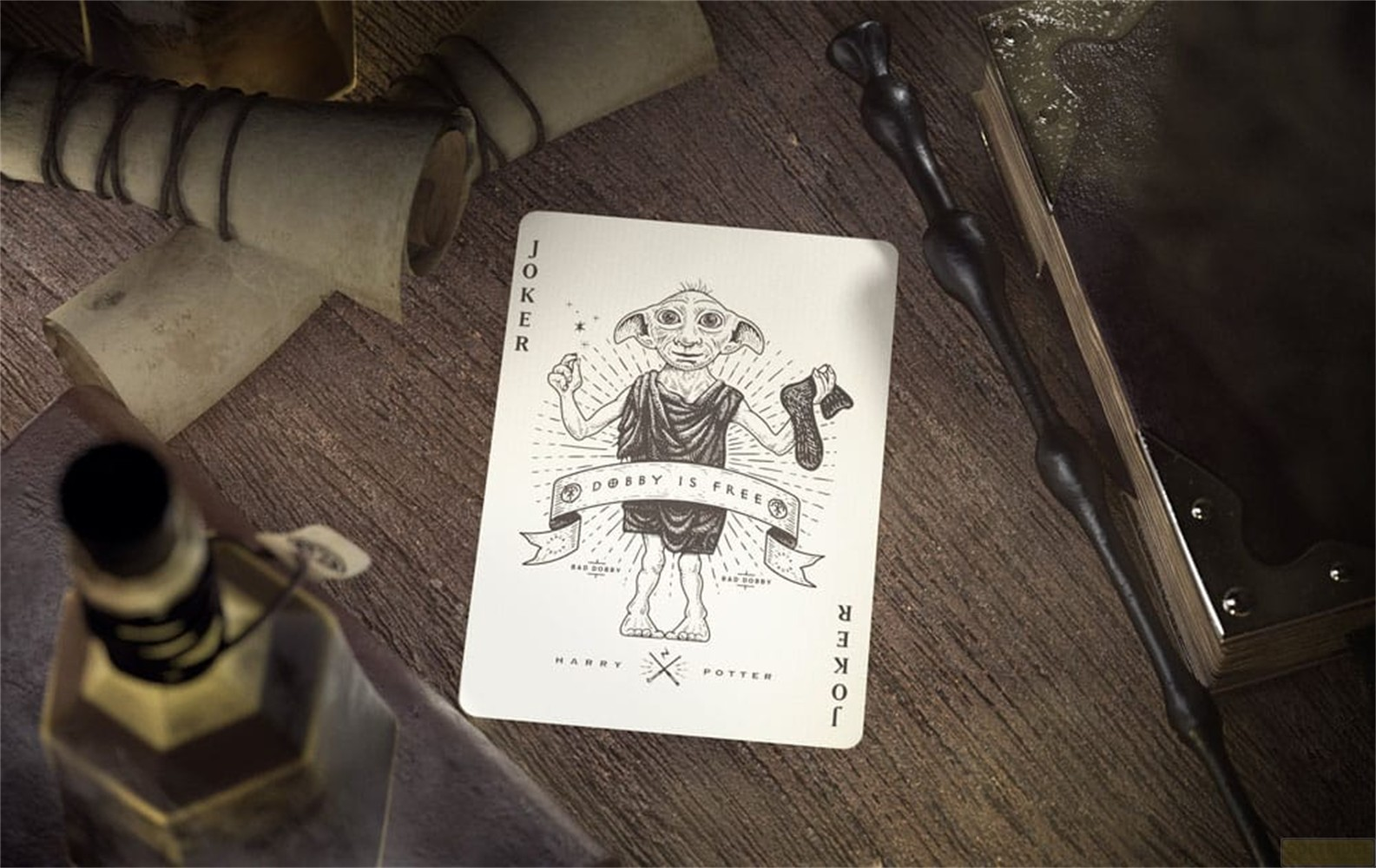Une carte Joker représentant Dobby de Harry Potter sur une table en bois avec des parchemins, une baguette et une potion.
