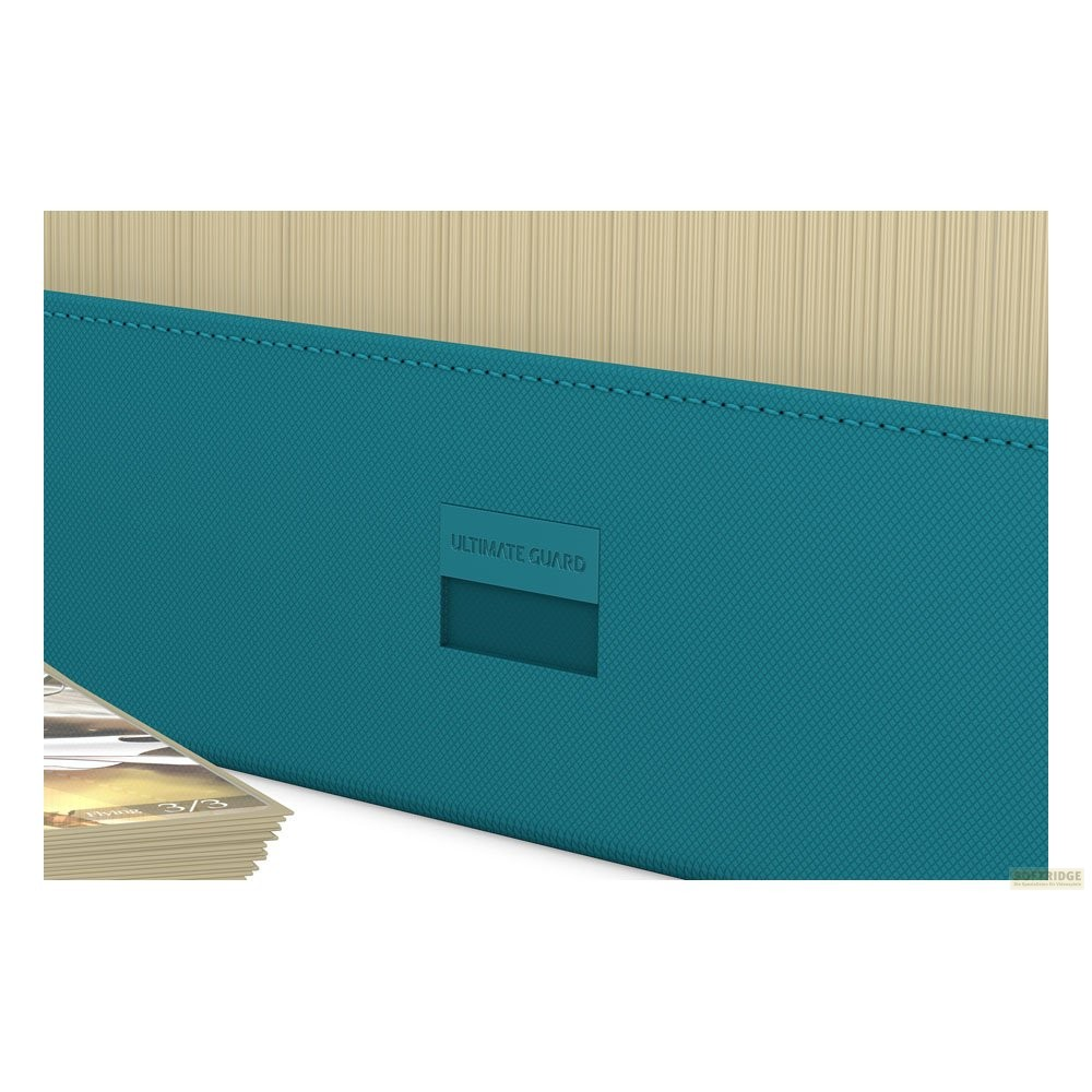Boîte de cartes Ultimate Guard turquoise avec coutures noires, une petite pile de cartes et un fond clair.