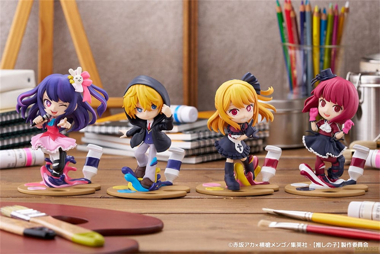 Cuatro figuras chibi coloridas de Oshi no Ko sobre bases de paleta, rodeadas de materiales de arte en un escritorio.