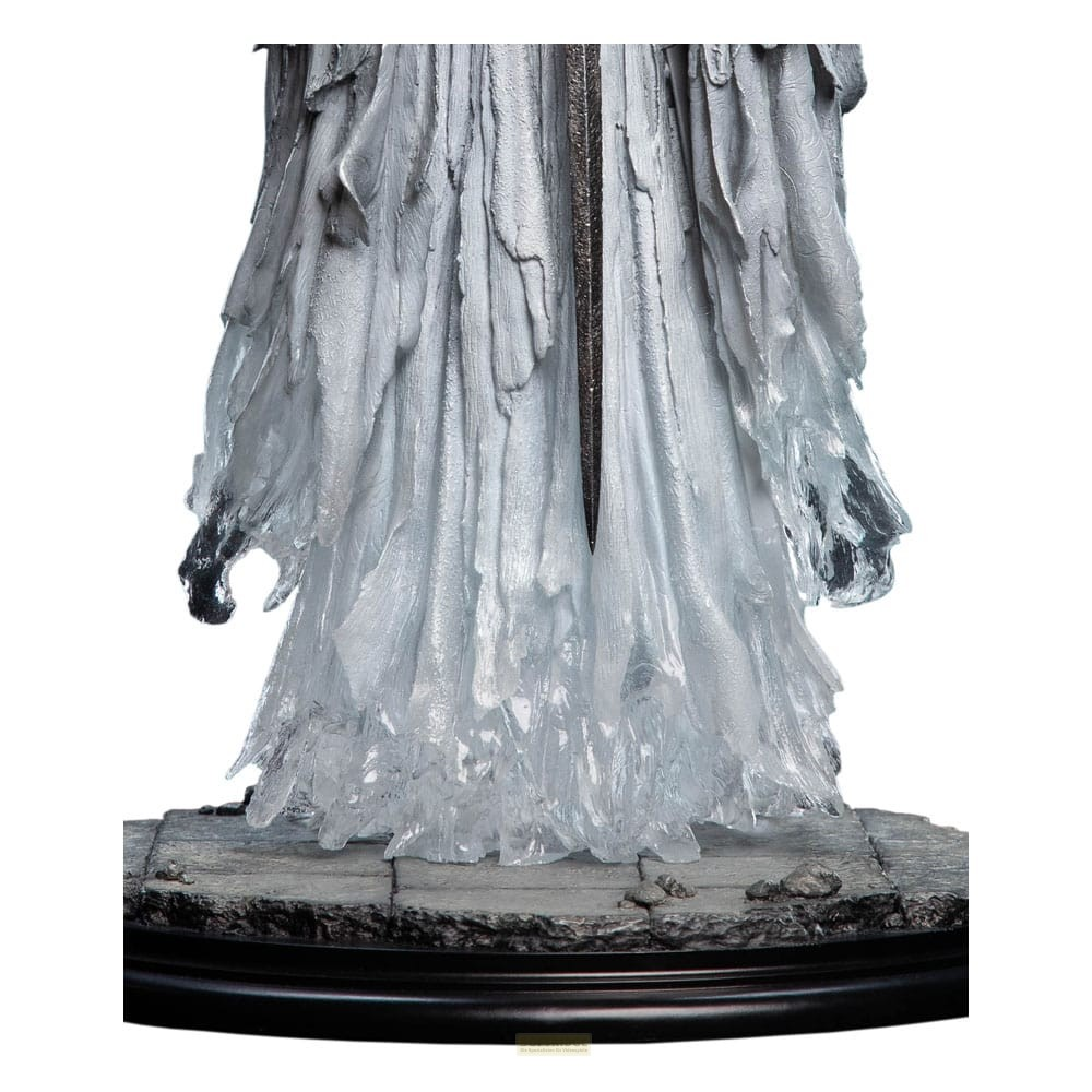 Una estatua gris de aspecto fantasmal con túnicas fluidas se alza sobre una base redonda y pedregosa contra un fondo blanco.