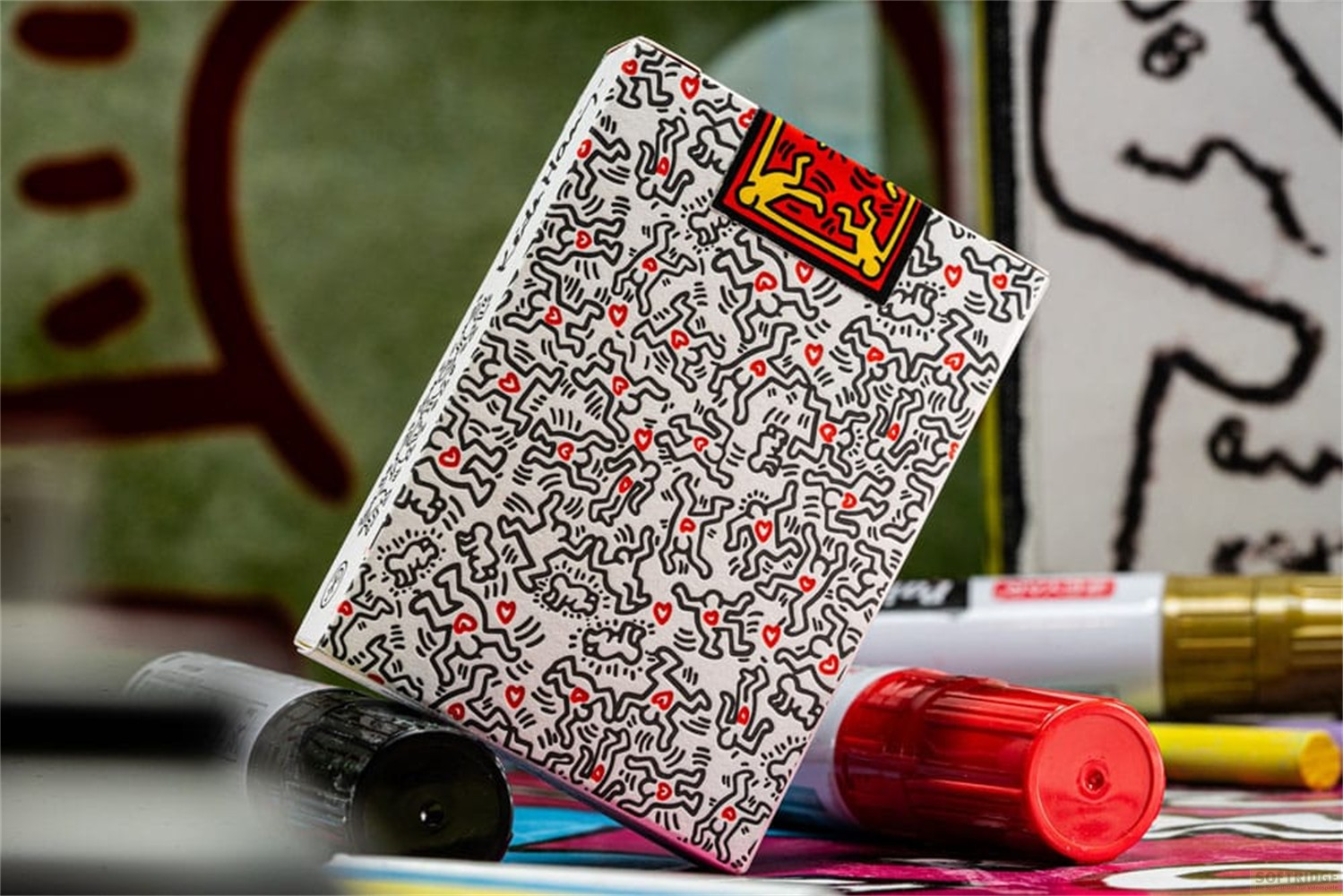 Une boîte de cartes à jouer style Keith Haring avec des danseurs et des cœurs rouges, entourée de marqueurs.