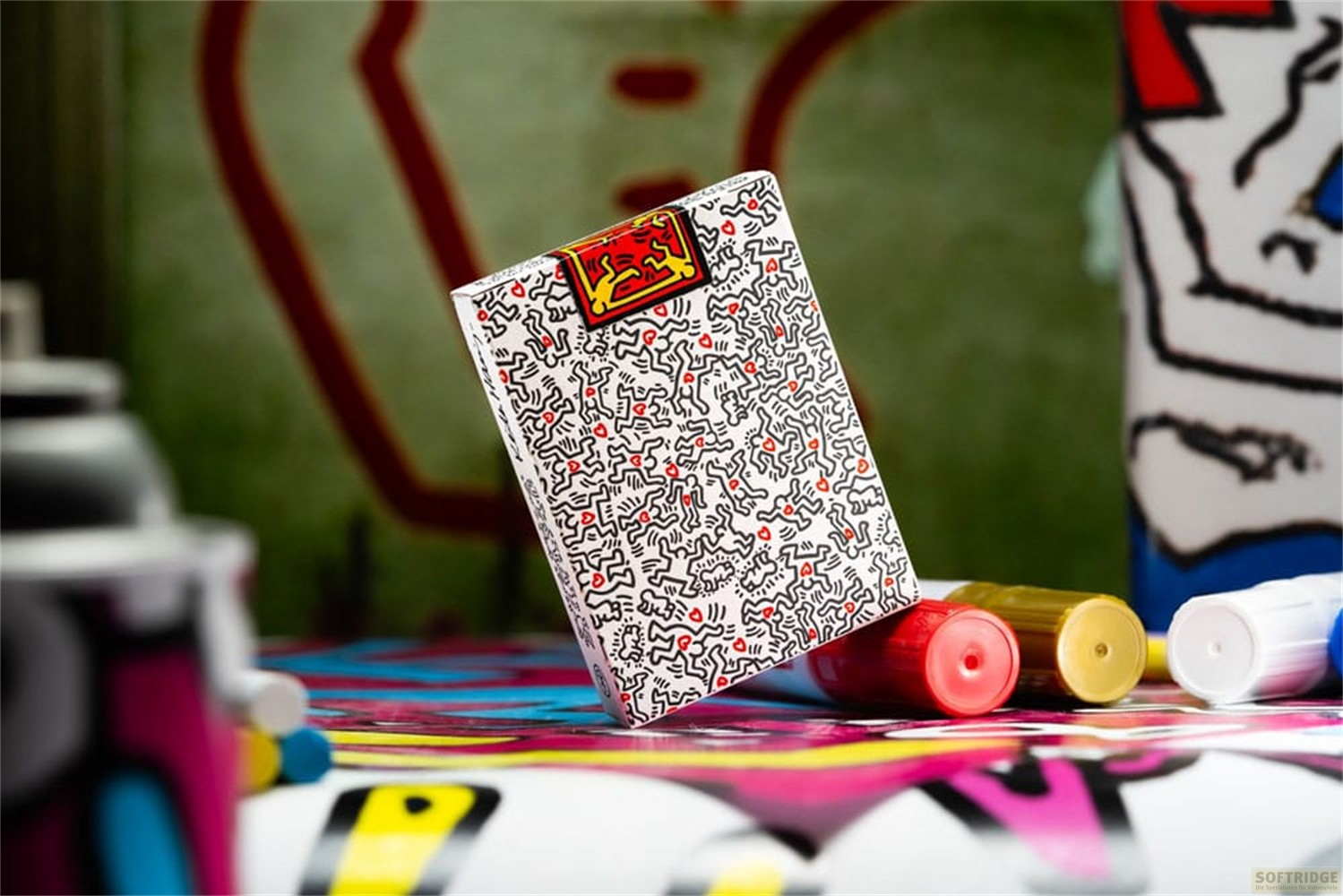 Un jeu de cartes au motif Keith Haring posé en diagonale sur une surface colorée style graffiti avec des feutres.