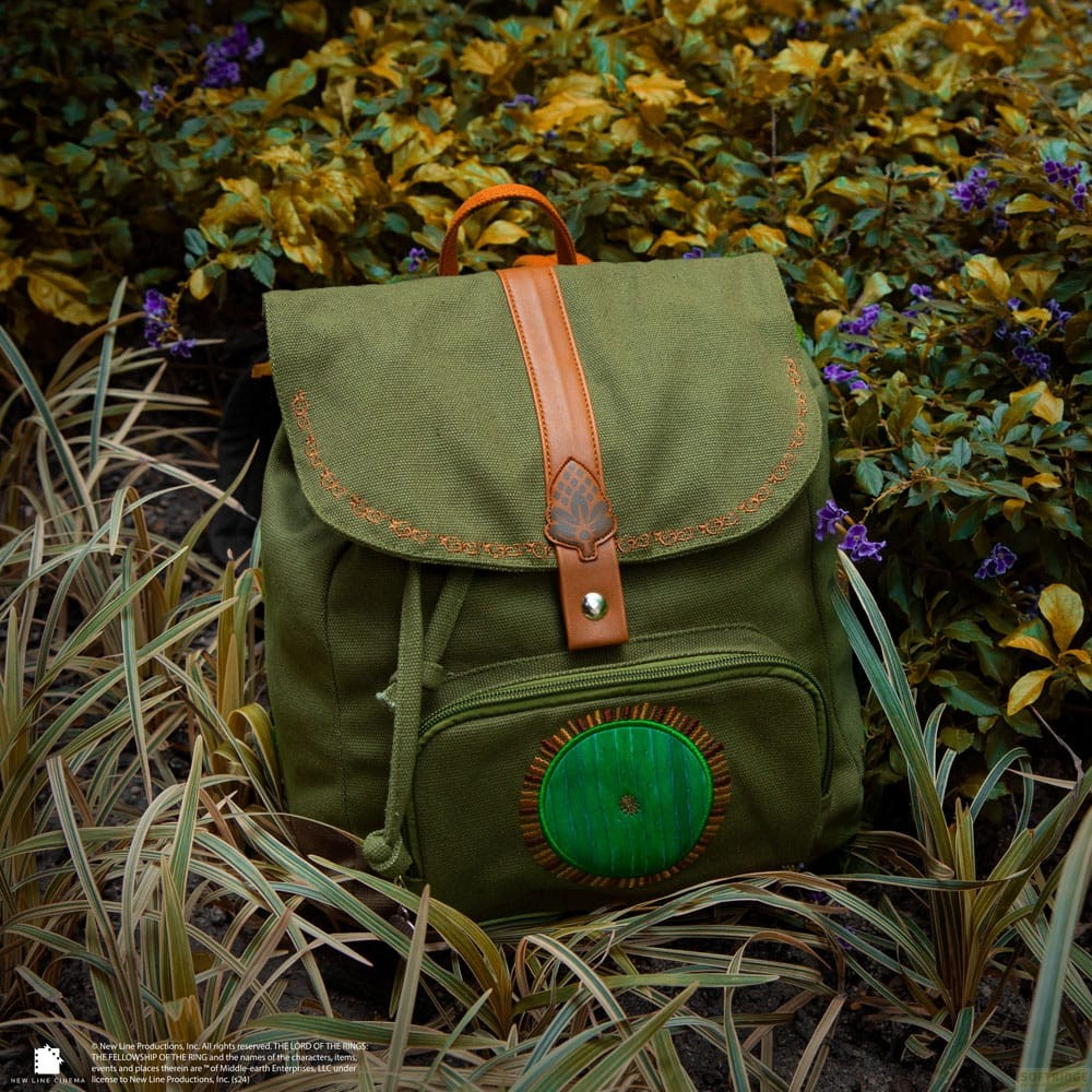 Una mochila verde con correas de cuero marrón y un detalle bordado redondo se encuentra entre arbustos y hierba alta.