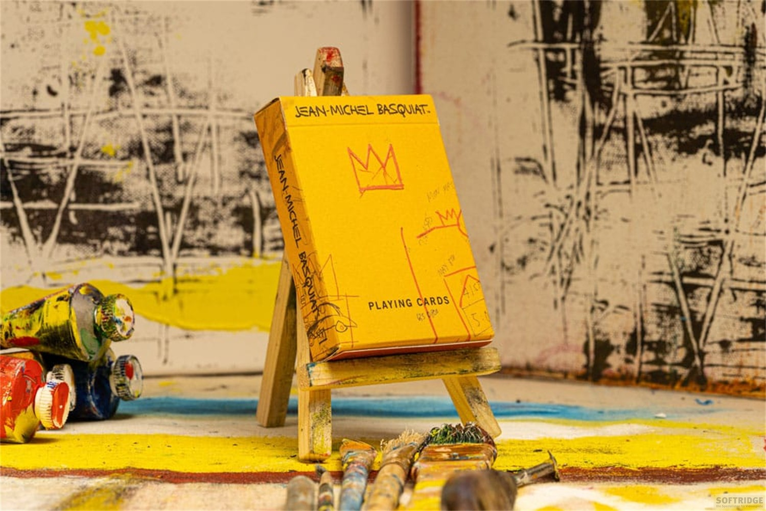 Boîte de cartes à jouer Jean-Michel Basquiat sur un mini chevalet en bois, entourée de tubes de peinture et de pinceaux.