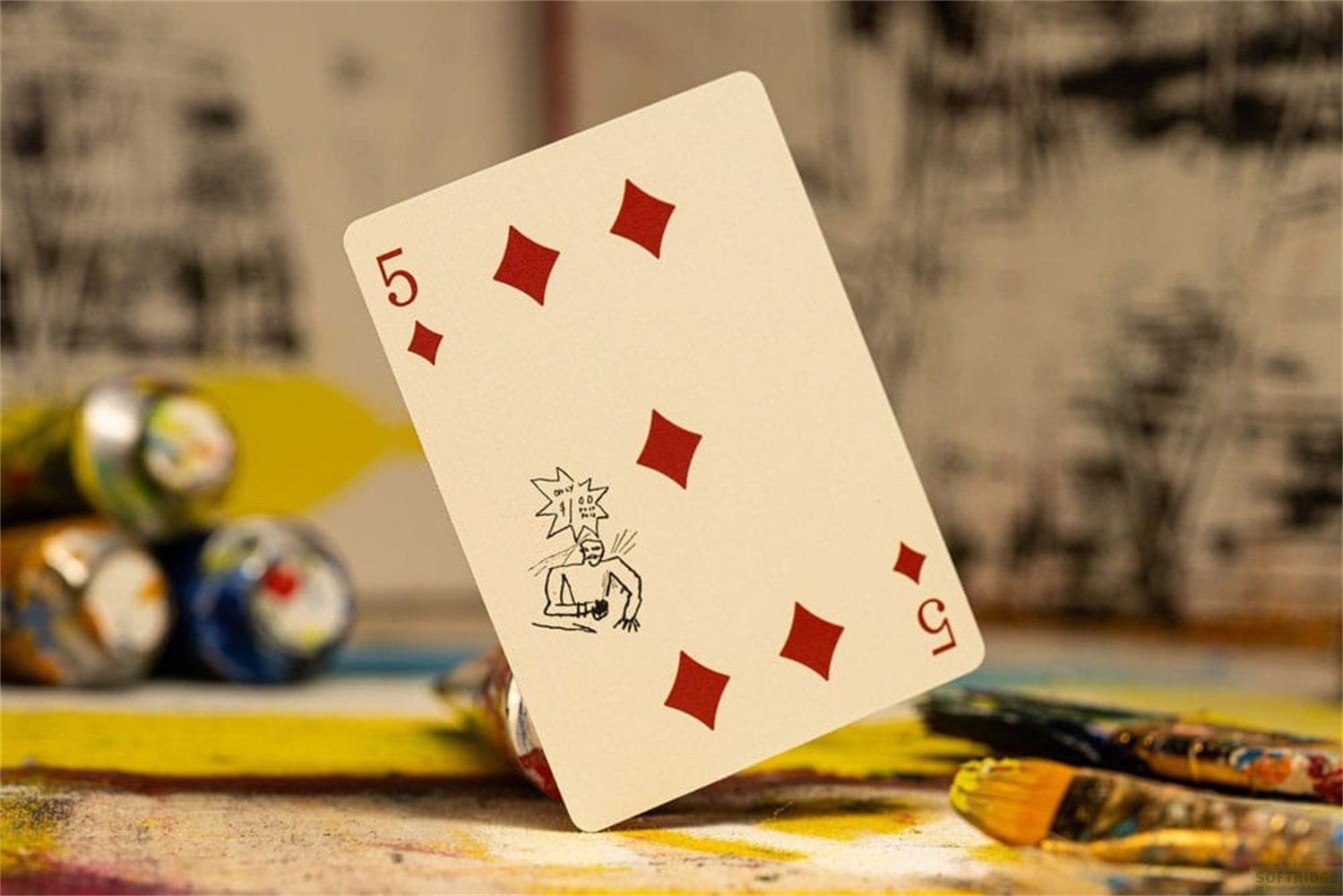 Une carte à jouer, le cinq de carreau, se dresse verticalement sur un tube de peinture au milieu de matériel artistique.