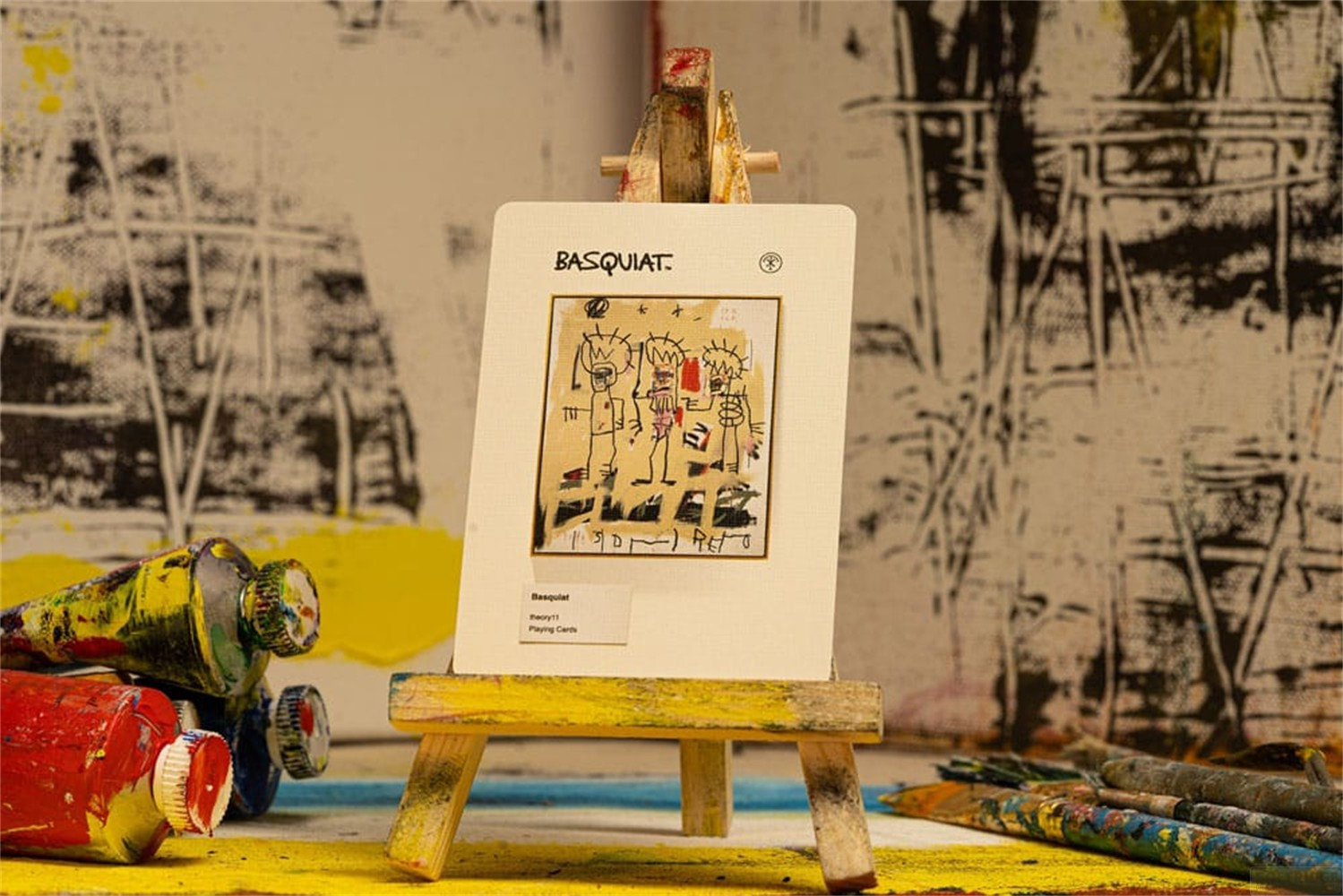 Une carte à jouer Basquiat sur un petit chevalet en bois, entourée de tubes de peinture et de pinceaux devant une toile.