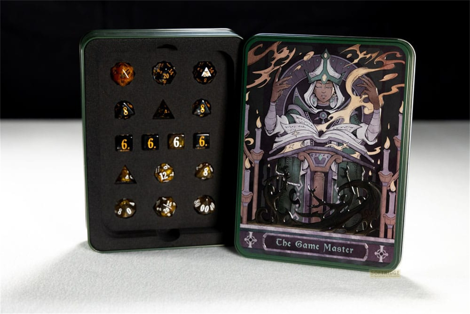 Une boîte en fer blanc ouverte affiche des dés noirs et dorés à gauche et l'art fantastique du Maître du Jeu à droite.