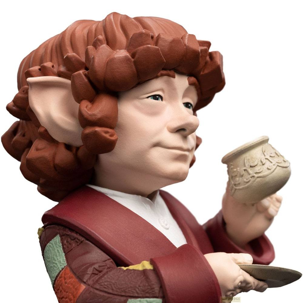 Una pequeña figura de hobbit con cabello castaño rojizo sostiene una taza y un platillo decorados sobre fondo blanco.
