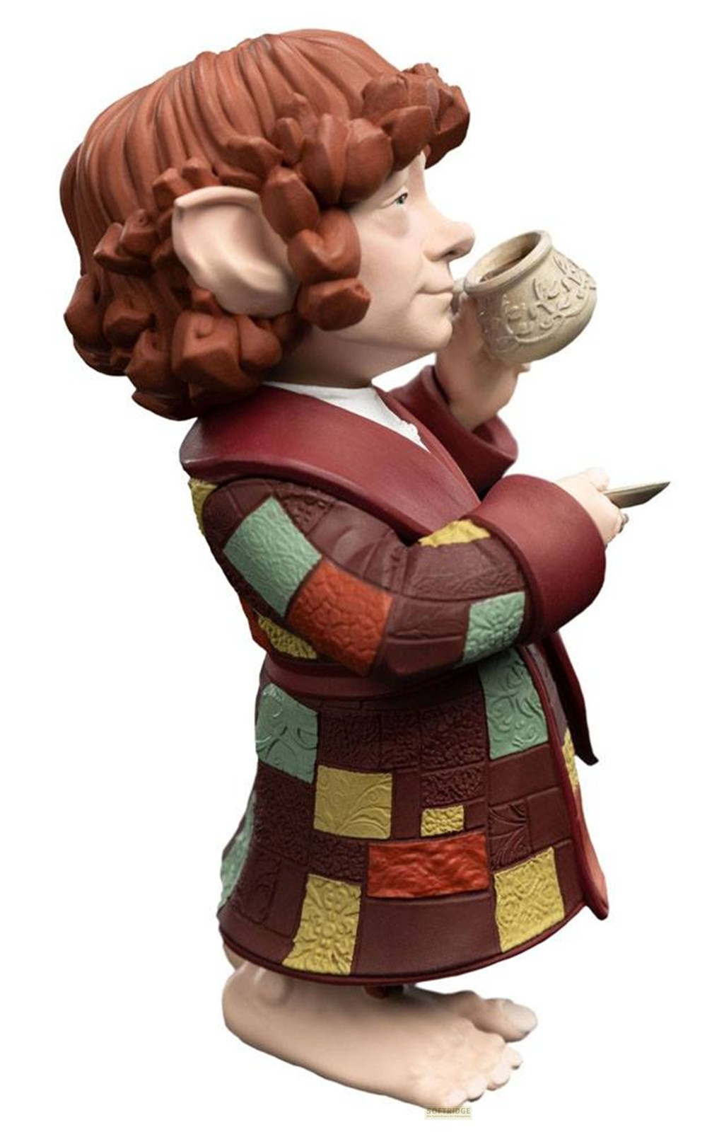 Figurina de un hobbit con cabello rojo vistiendo una bata de retazos colorida, bebiendo de una taza.