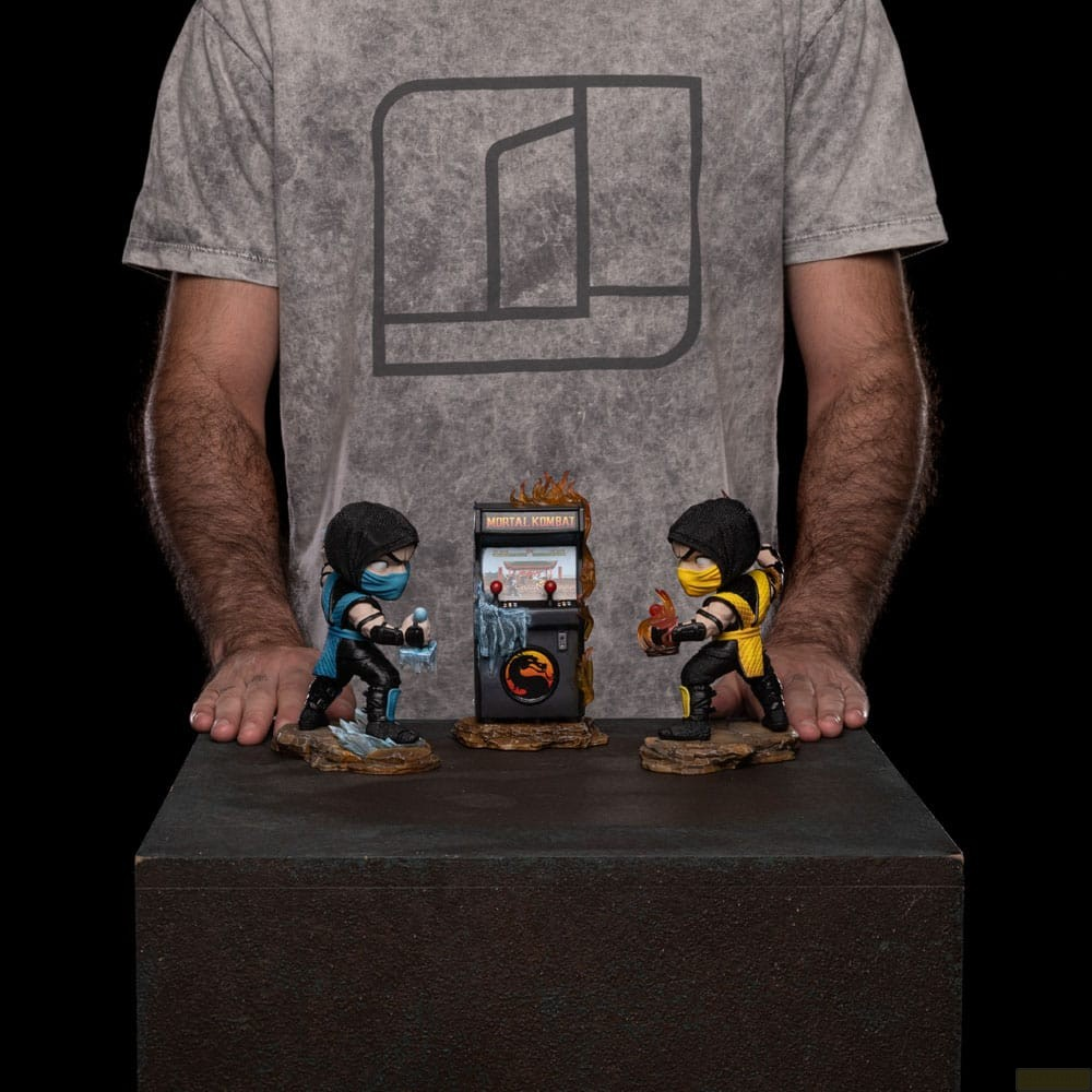 Una persona con camisa gris presenta dos figuras de Mortal Kombat junto a una minigabinete arcade.