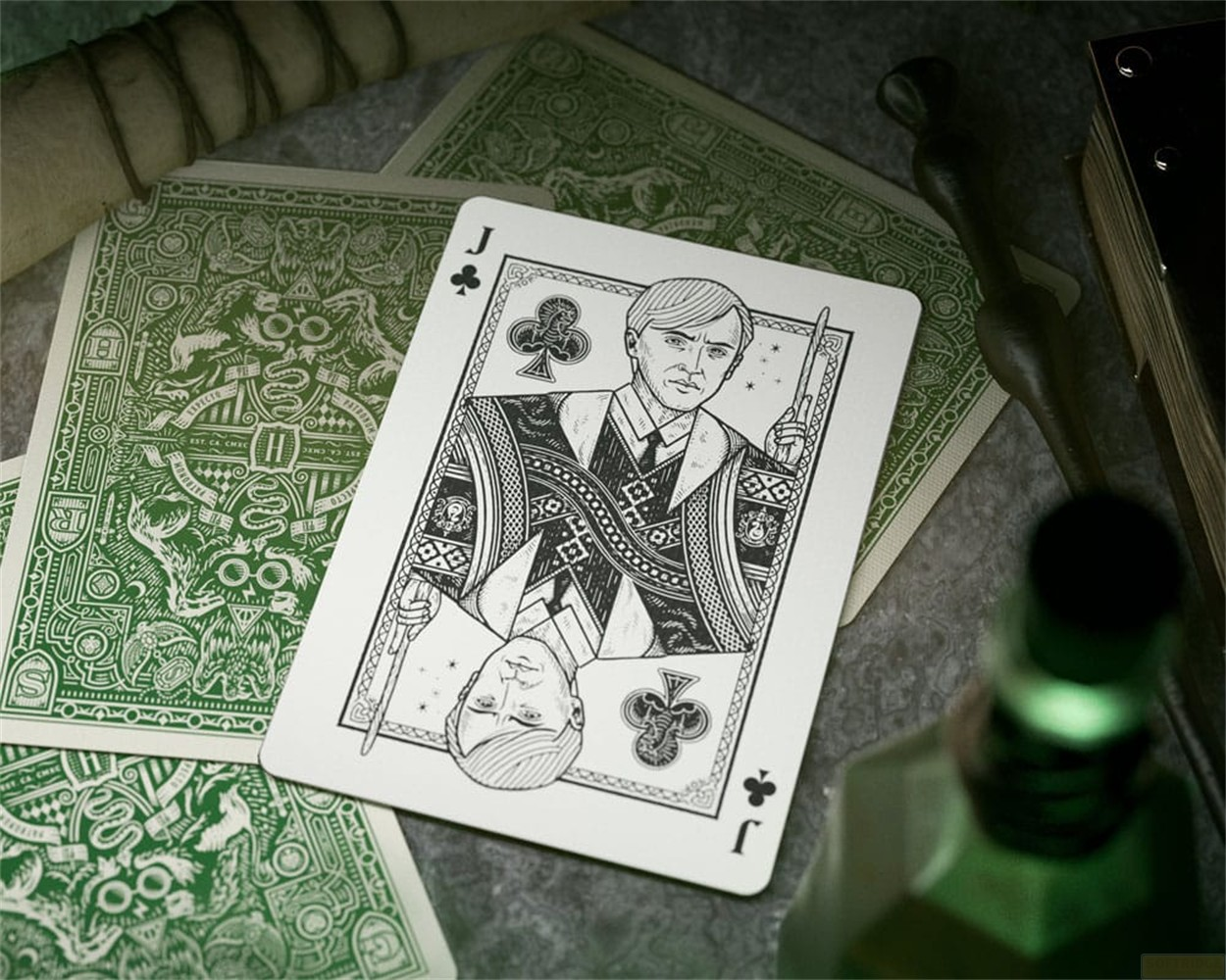 Une carte de valet de trèfle repose sur une table parmi des cartes vertes à motifs, une baguette, un parchemin et une potion.