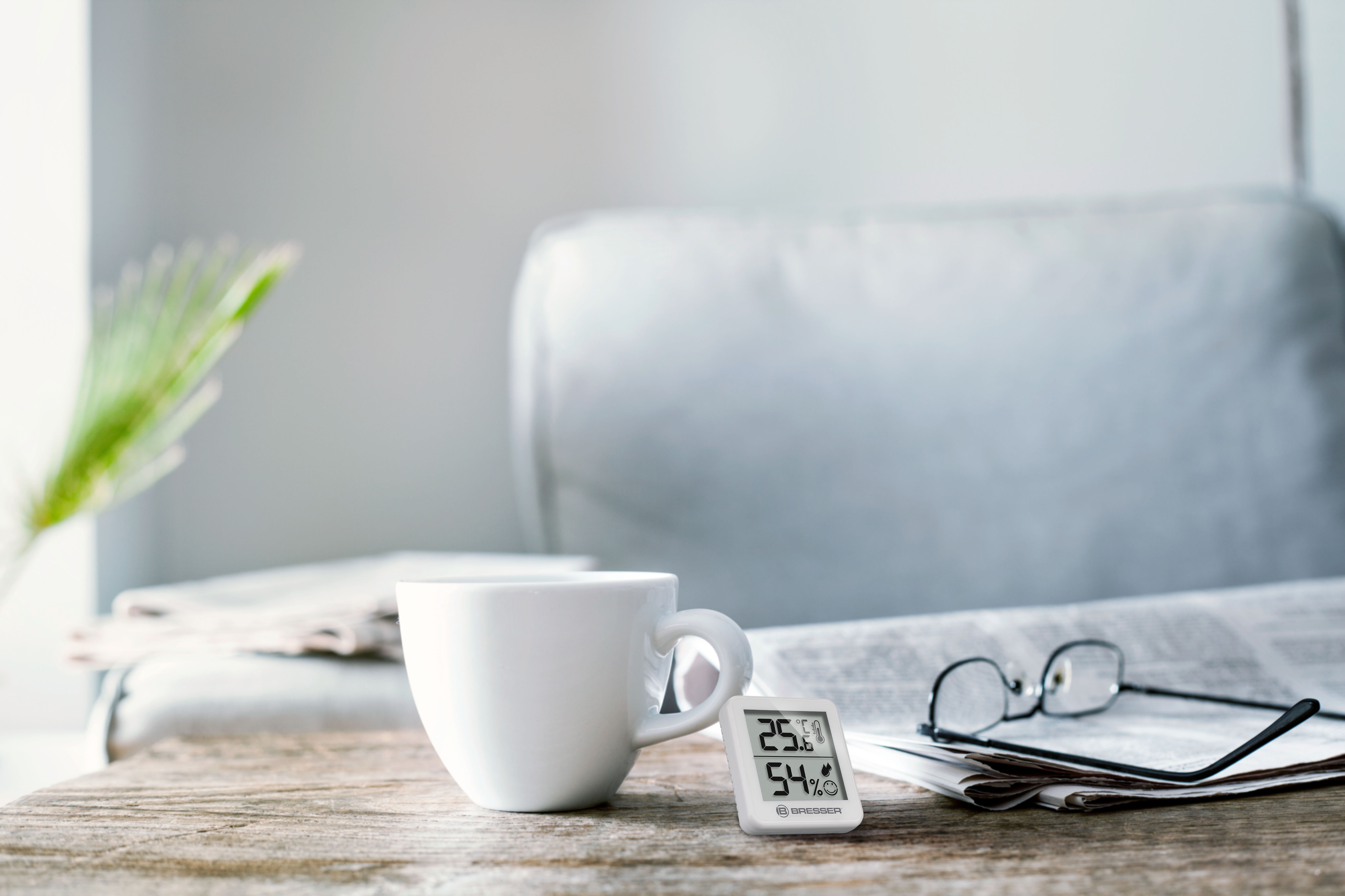 Una taza blanca, gafas y un pequeño termómetro en una mesa de madera frente a un sillón gris.