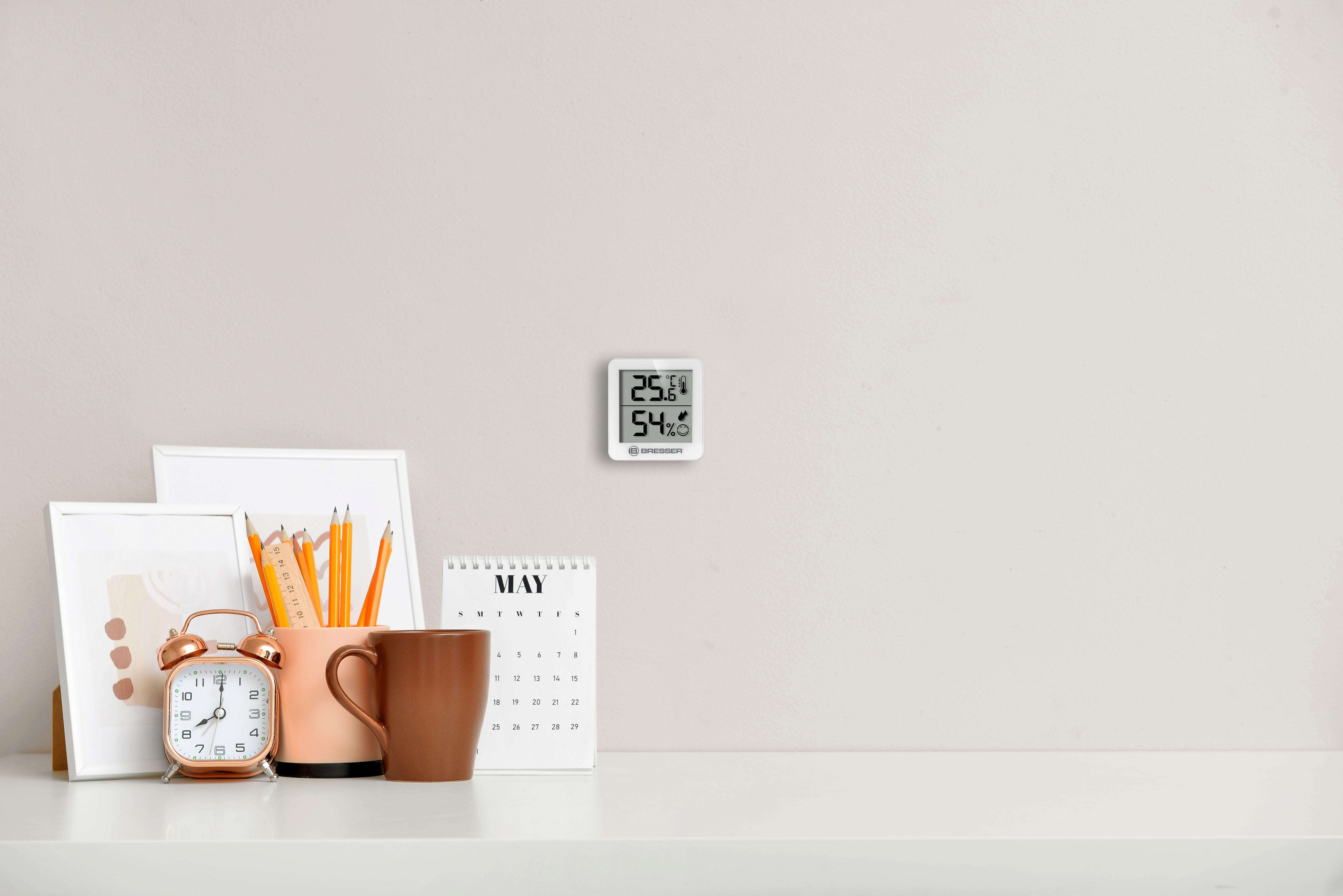 Un escritorio blanco con termómetro de pared, reloj, lápices y calendario de mayo.