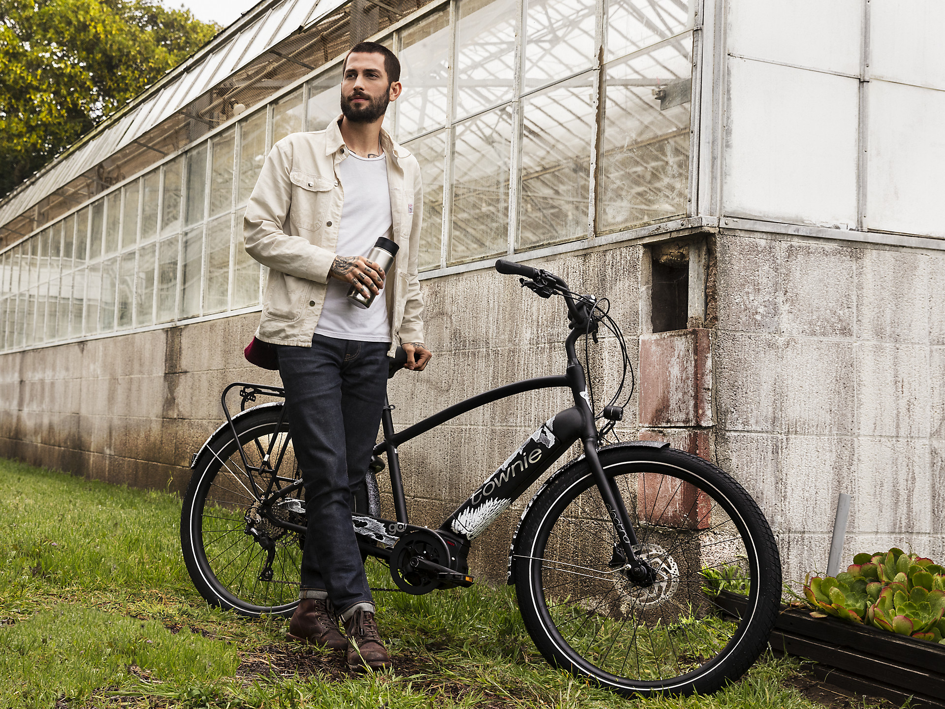 Ein Mann steht draußen neben einem schwarzen E-Bike, hält eine Flasche, im Hintergrund ein Gewächshaus.