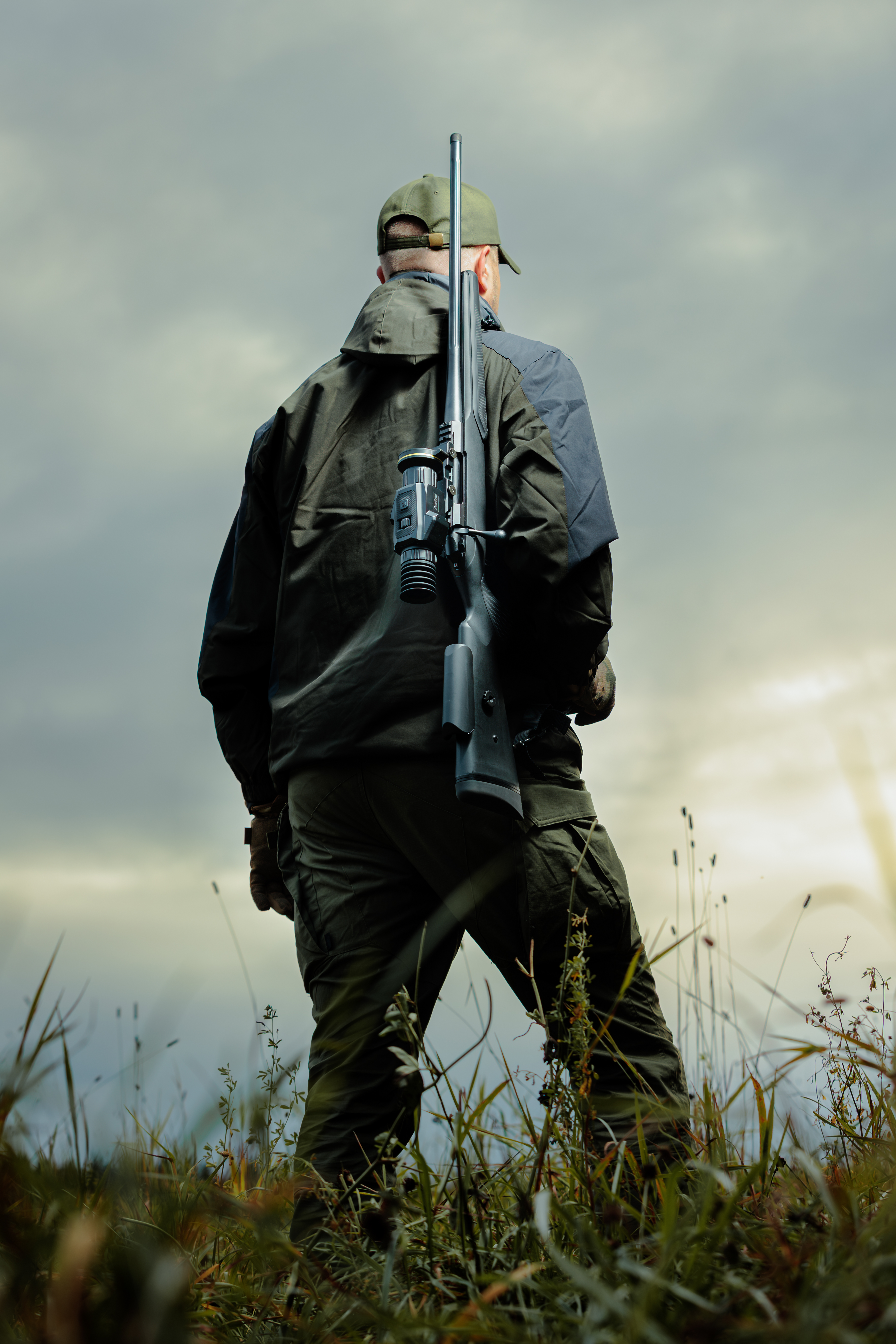 Un cazador con ropa de camuflaje y gorra está de pie en hierba alta, sosteniendo un rifle.