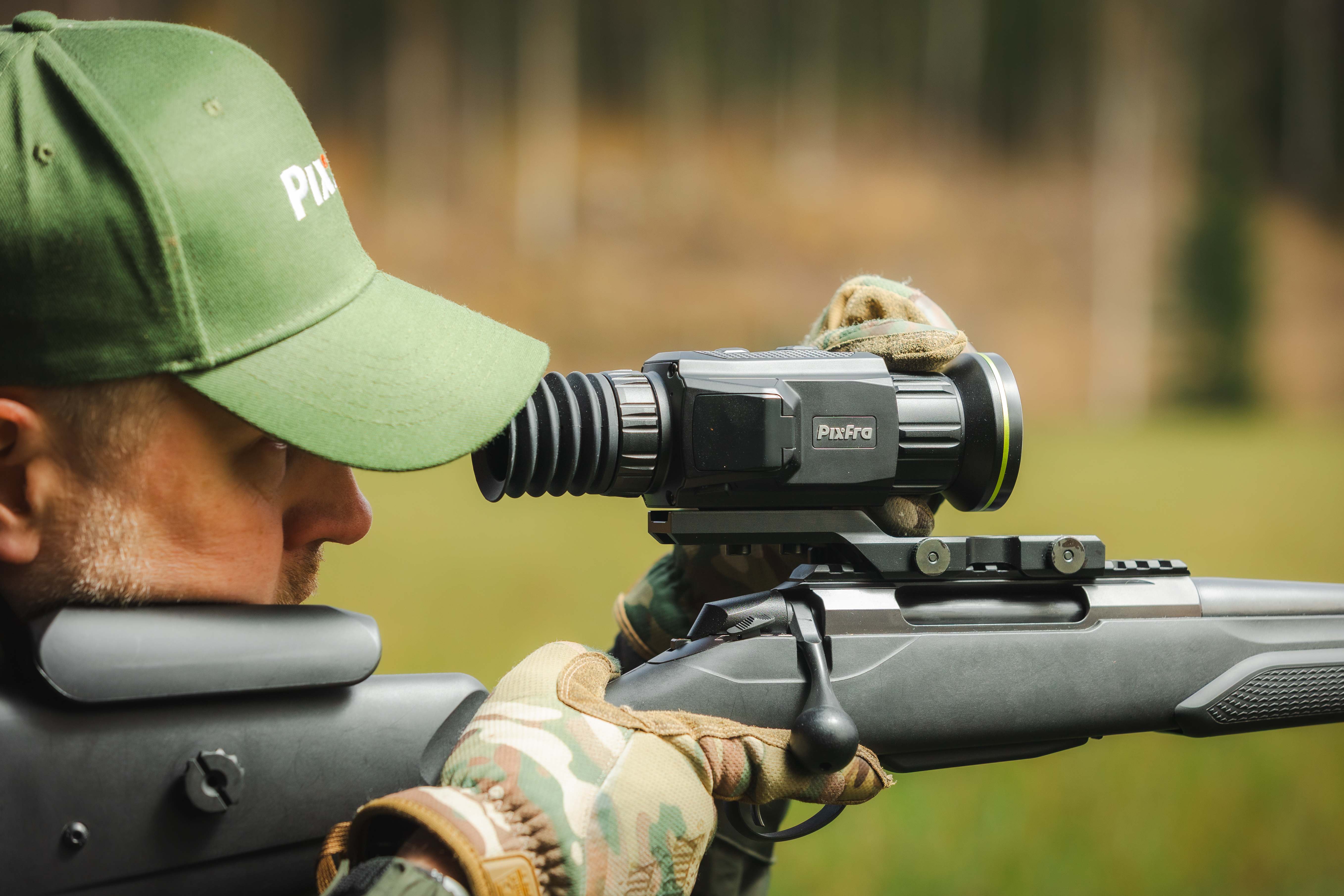 Un cazador con gorra verde y guantes de camuflaje apunta con un rifle con visor nocturno en un bosque.