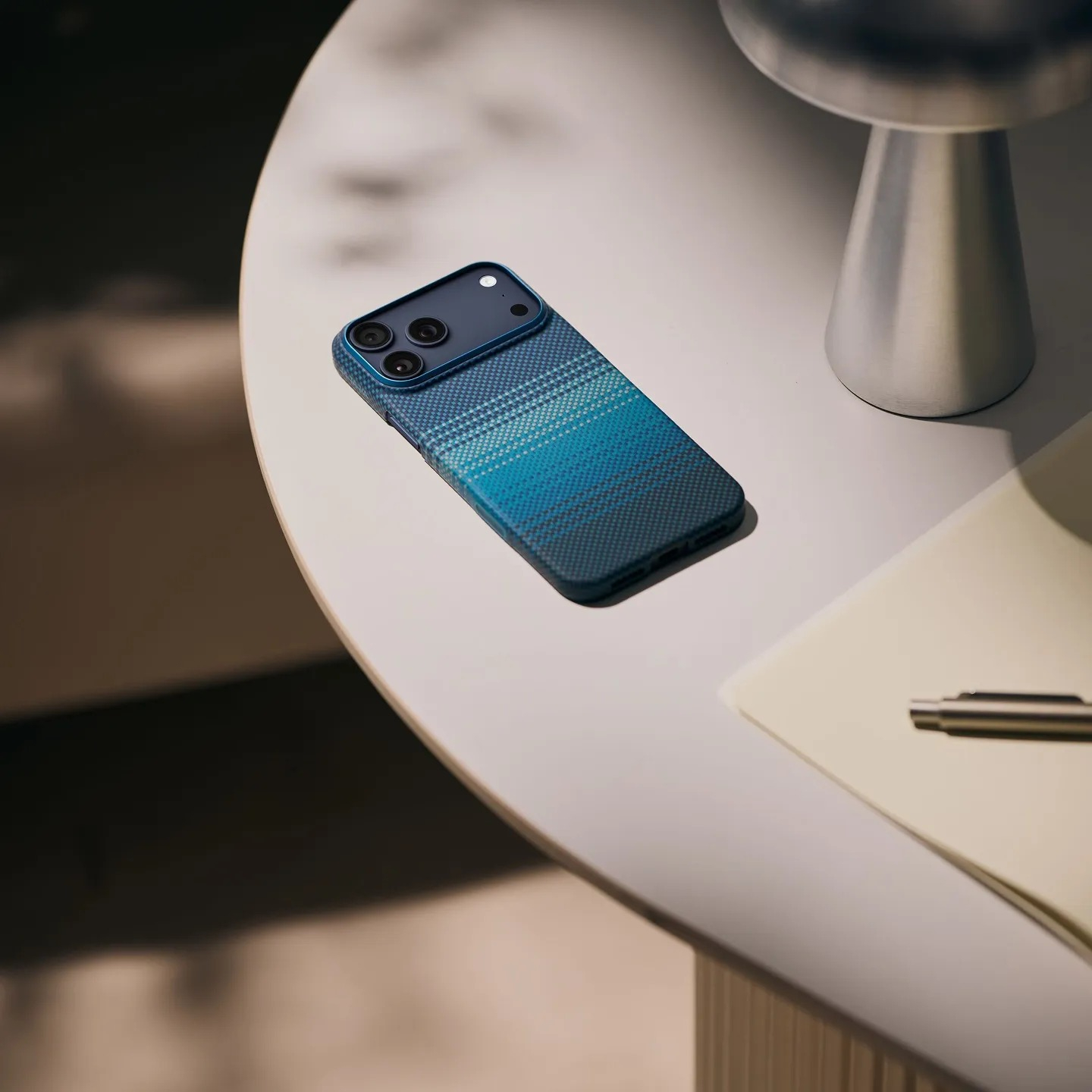 Niebieski smartfon w wzorzystym etui na białym stole obok lampy i notesu.