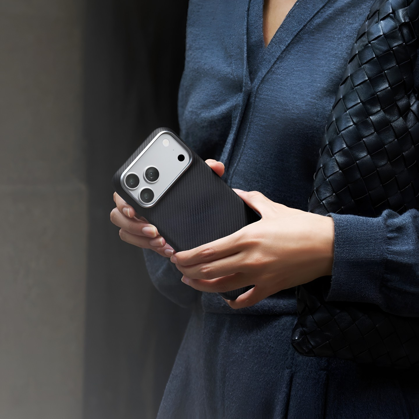Kobieta w niebieskim topie trzyma smartfon w czarnym etui i niesie czarną skórzaną torbę.
