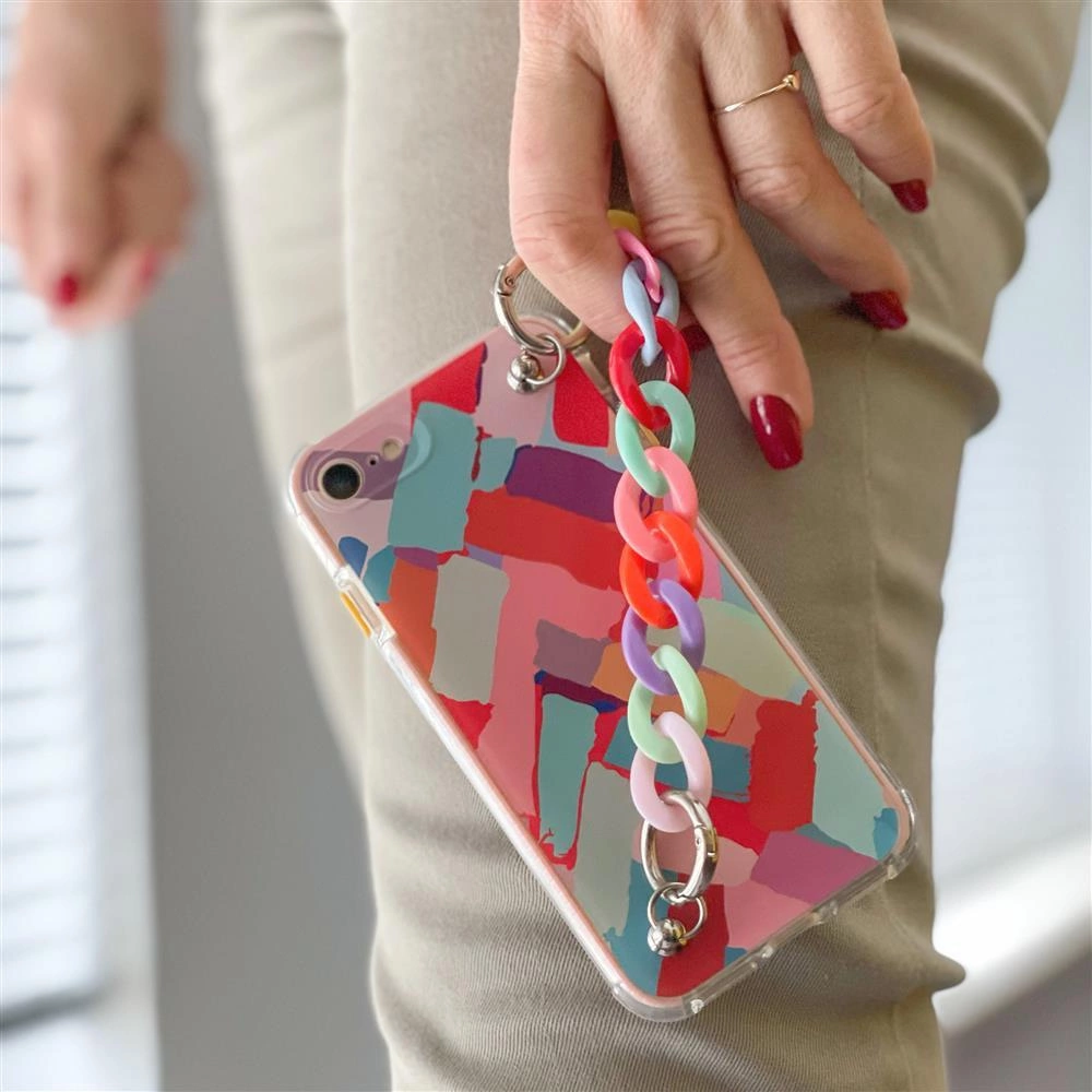 Smartphone in custodia astratta colorata con catena pastello, tenuto da una donna in pantaloni beige.