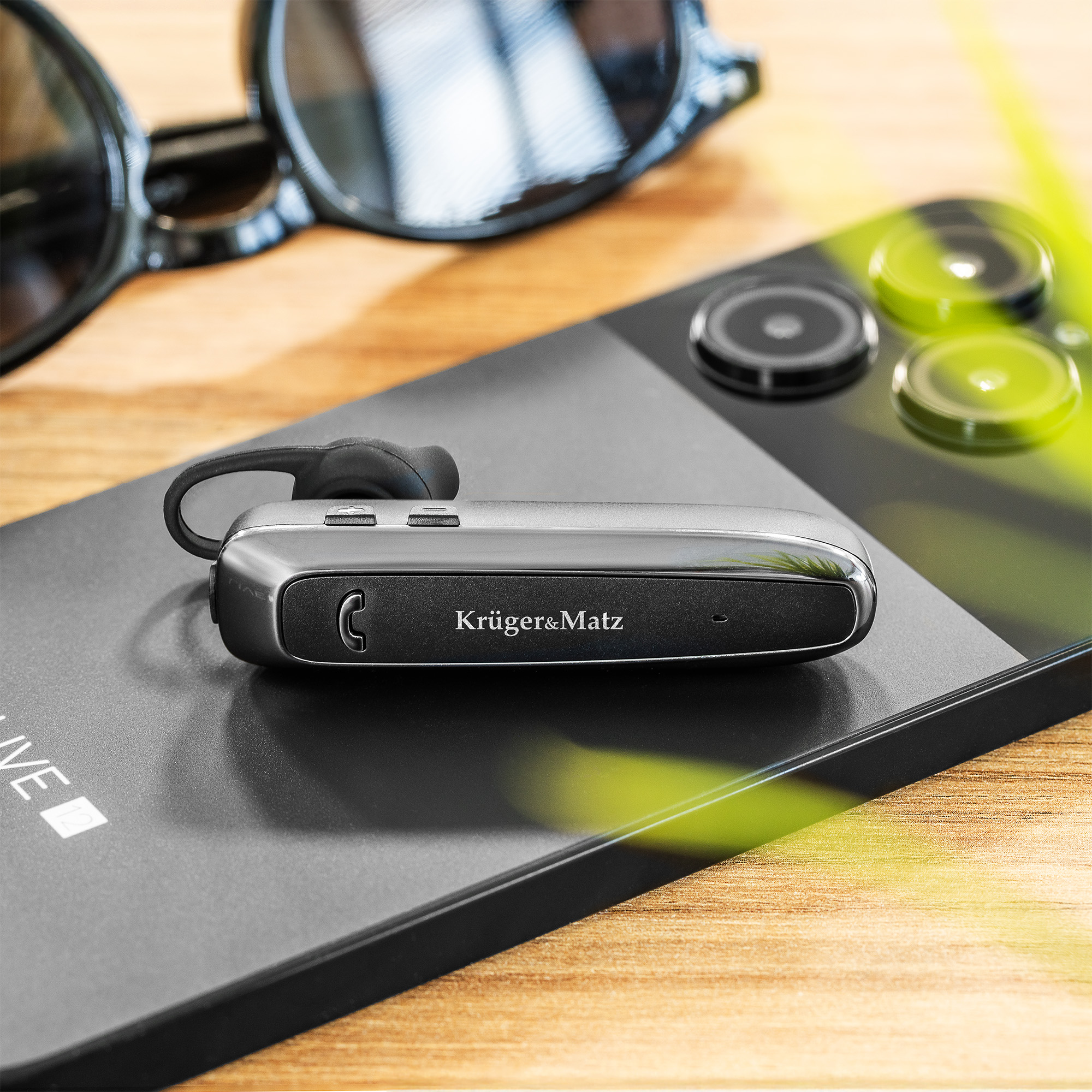 Sluchawka Bluetooth Kruger&Matz lezy na smartfonie obok okularów przeciwslonecznych.