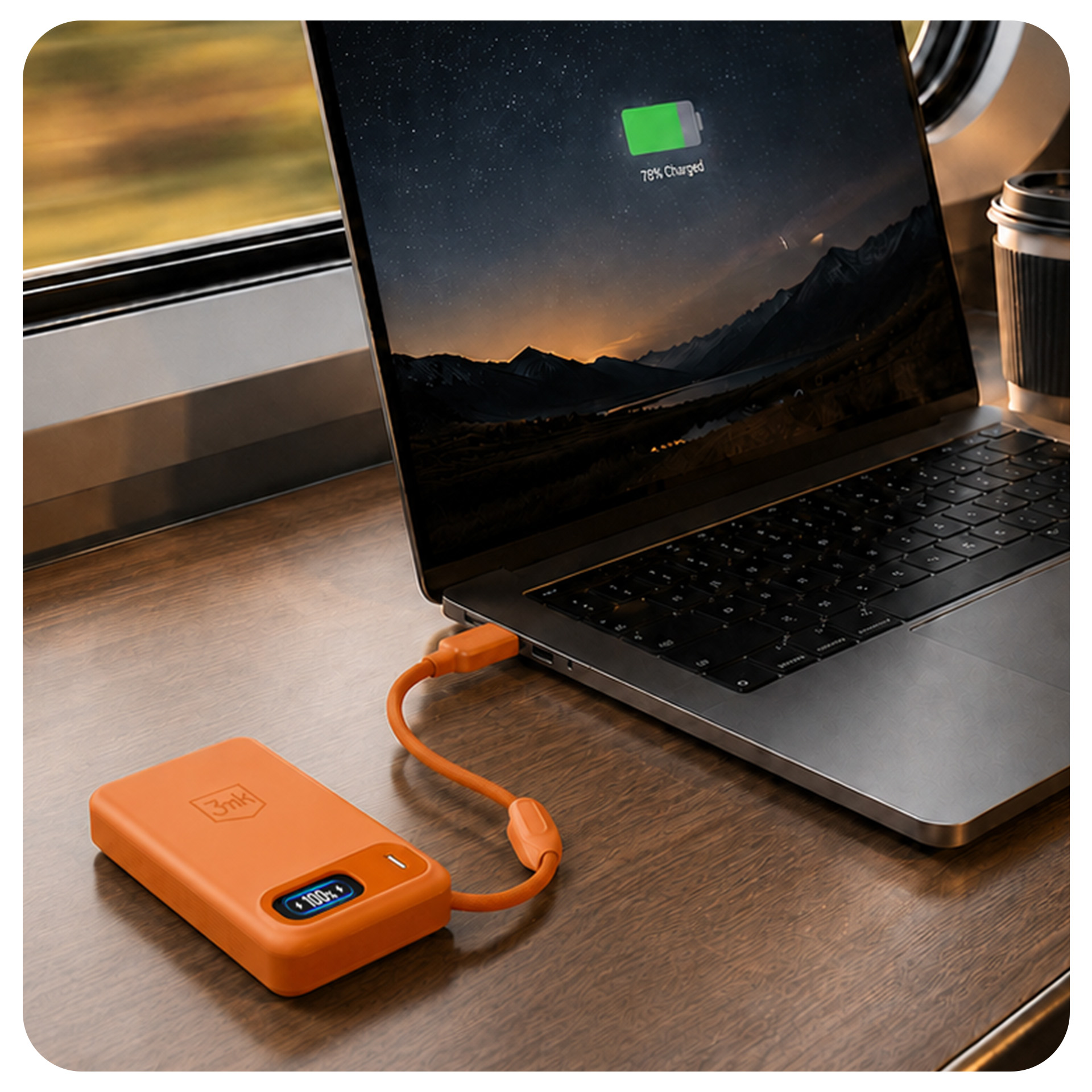 Pomarańczowy powerbank ładuje laptopa na drewnianym stole przy oknie.