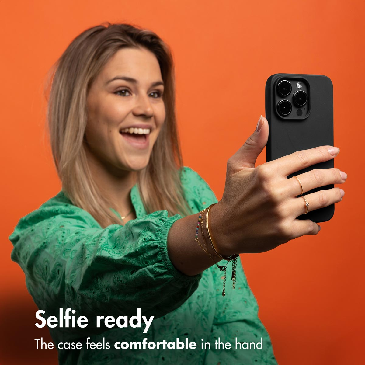 Uśmiechnięta blondynka w zielonym topie trzyma czarny smartfon na pomarańczowym tle.