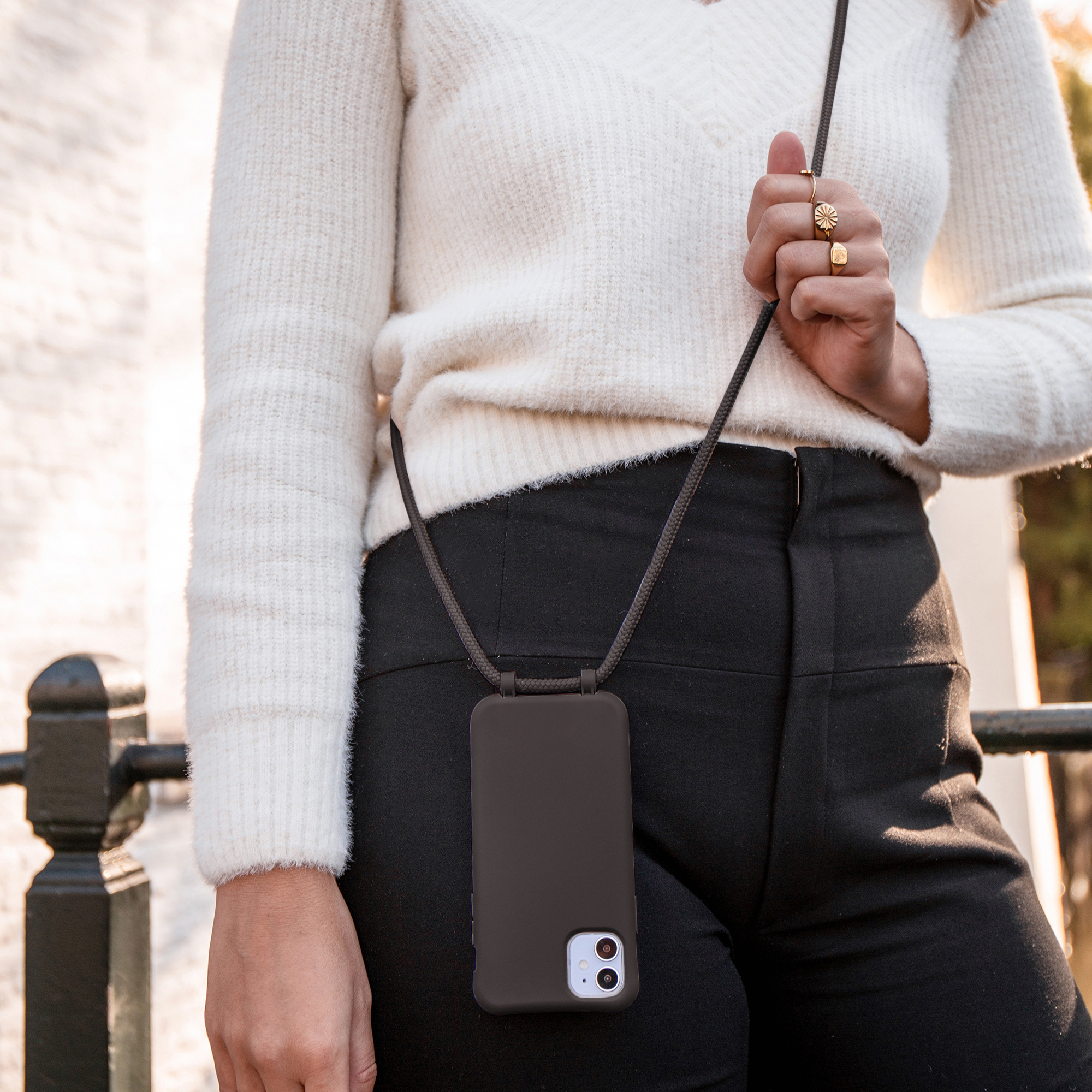 Une femme en pull blanc porte un smartphone noir avec bandoulière.