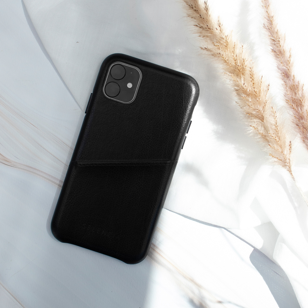 Ein schwarzes Leder-Smartphone-Etui mit integrierter Tasche liegt auf weißem Stoff neben weichen, trockenen Blumen.